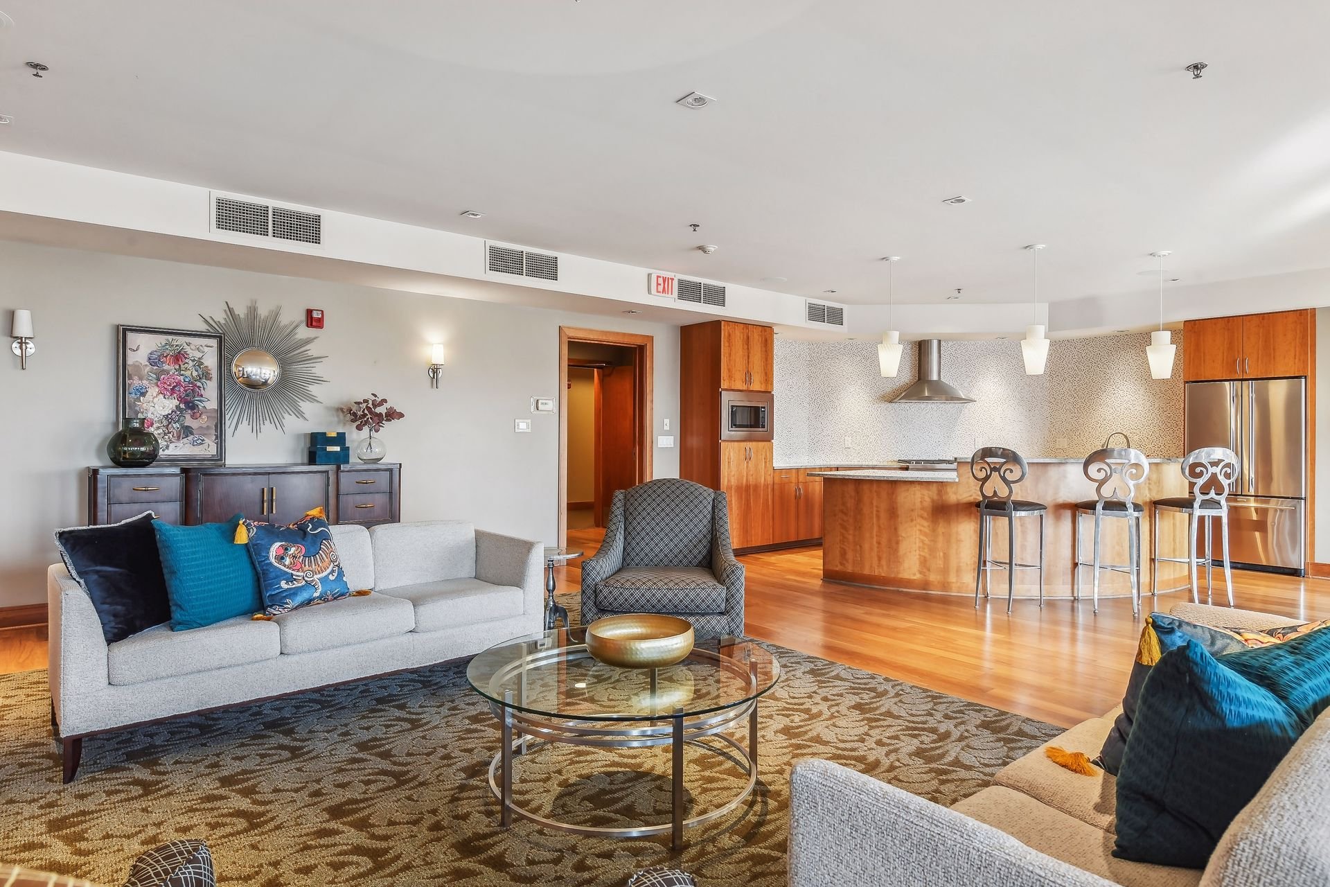 Community room and kitchen with beige sofas, dark wood sideboard with wall art, patterned rug, and kitchen island with bar stools, stainless steel appliances, and hanging pendant lights.