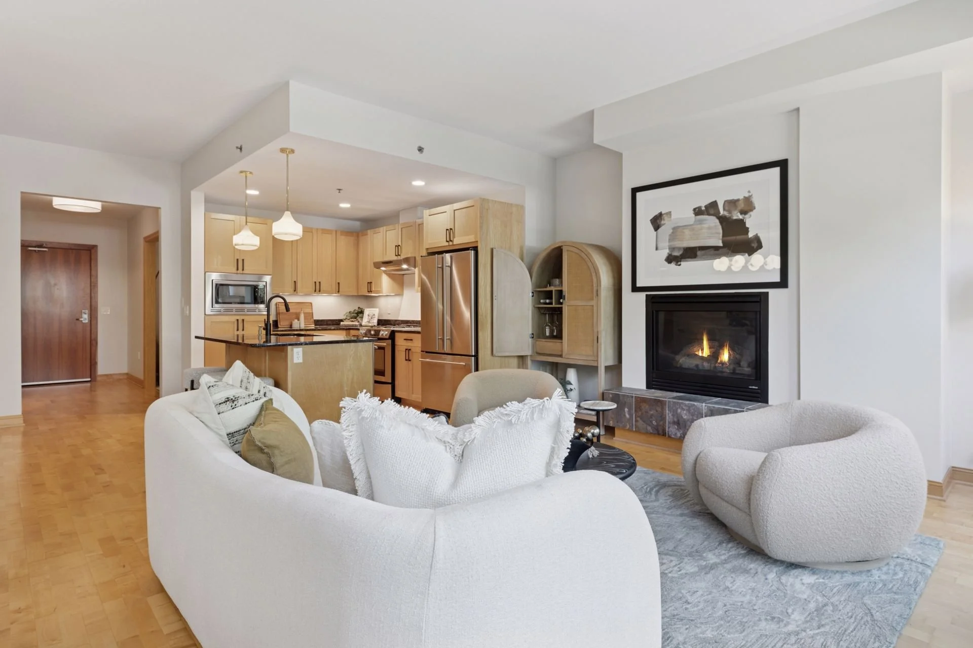Modern living room with fireplace and open kitchen in background with wooden cabinets and pendant lighting.