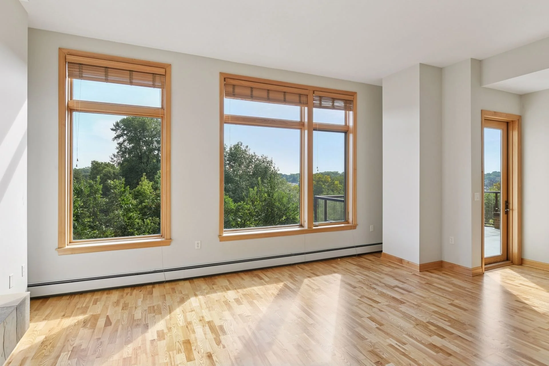 Empty living room with large windows and a glass door, hardwood floors, white walls, and wooden window and door frames.