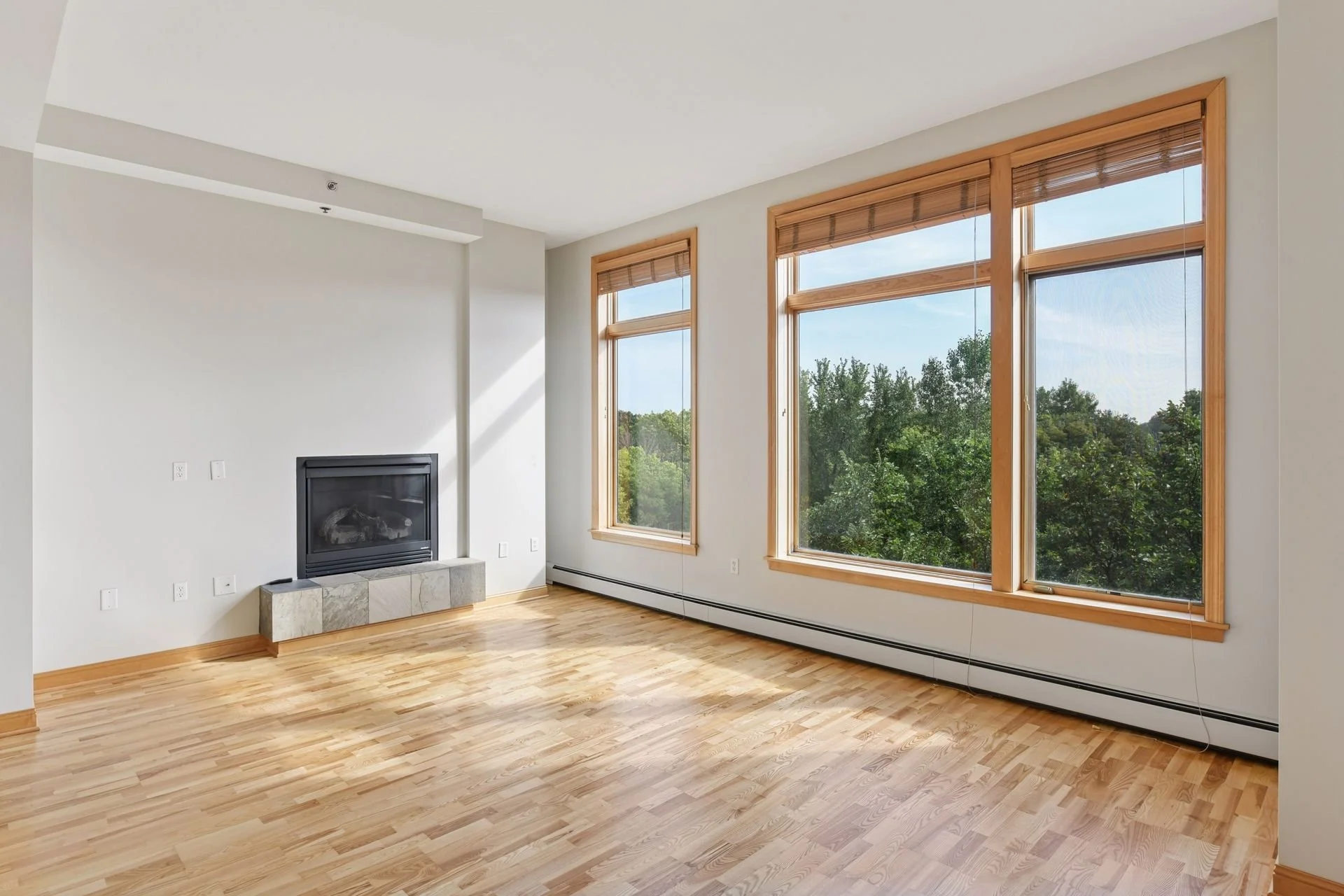 Empty living room with large windows, hardwood flooring, white walls, a fireplace, and wood trim.