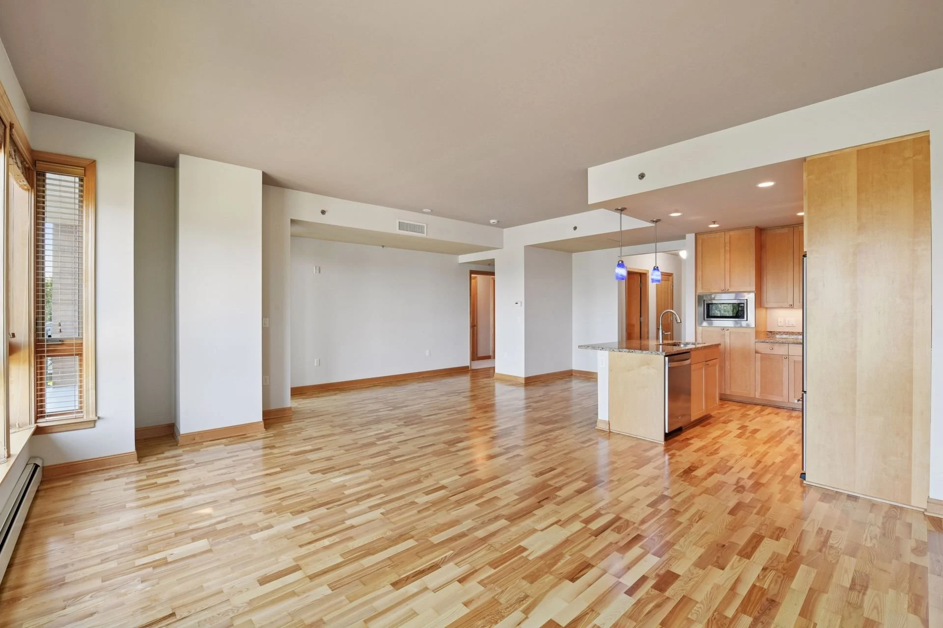 Living area with hardwood floors, large windows with blinds, and an open kitchen with a small island and wooden cabinetry.