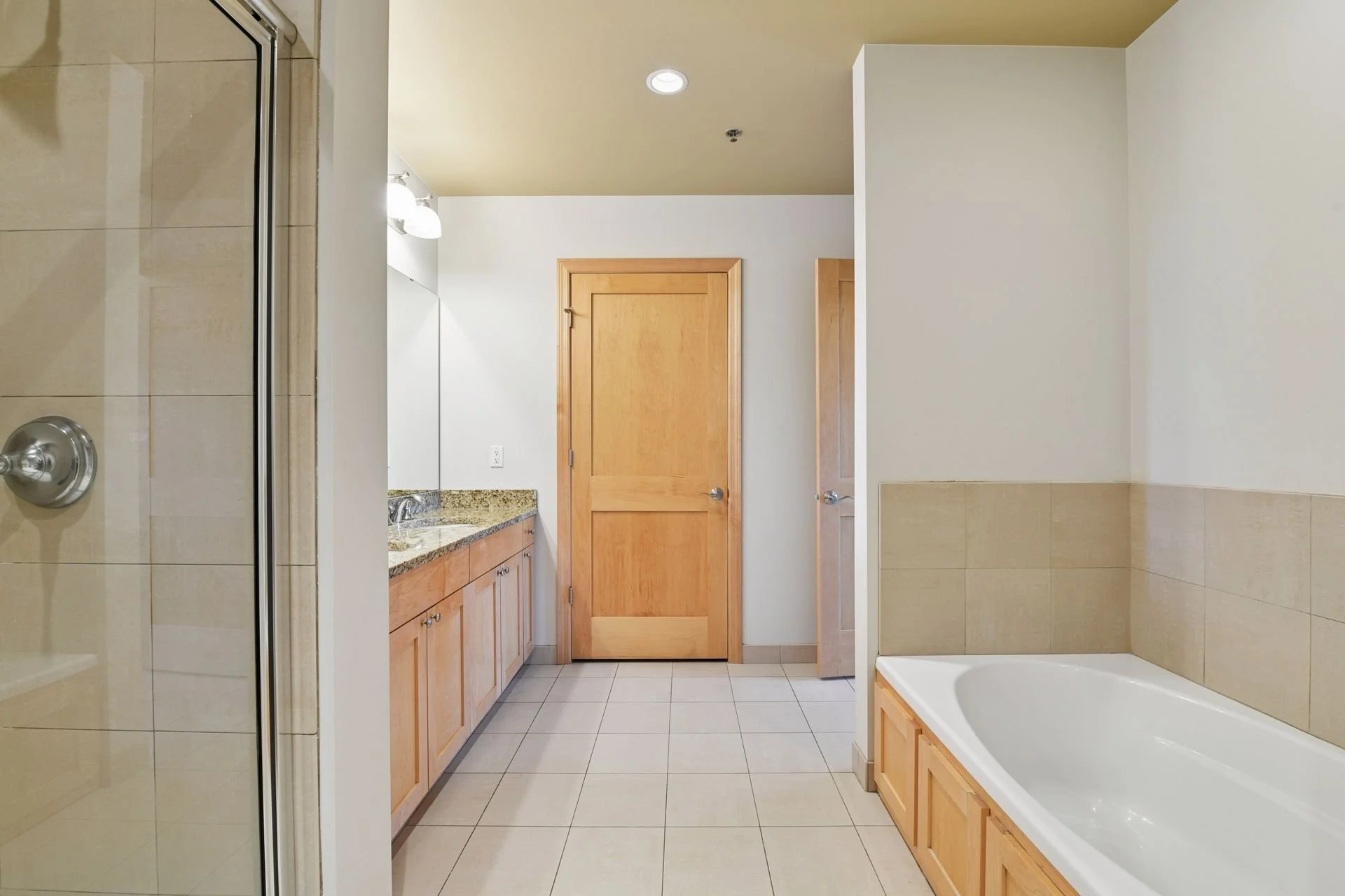 Bathroom with shower on the left, wooden vanity with granite countertop and large mirror in the middle, and bathtub on the right, all with tiled walls and floor.