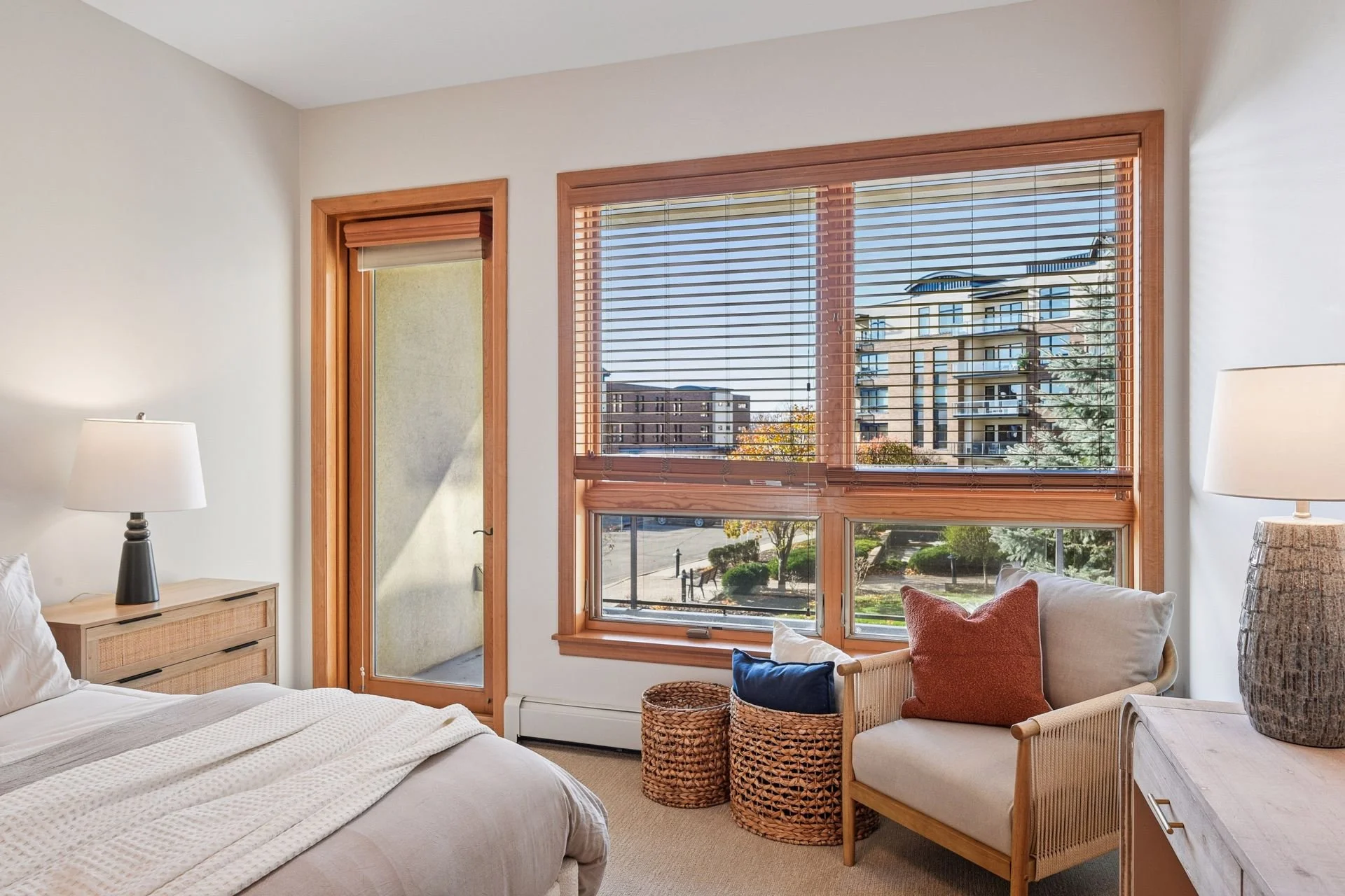 A bedroom with large windows, revealing an outdoor view of buildings and trees.