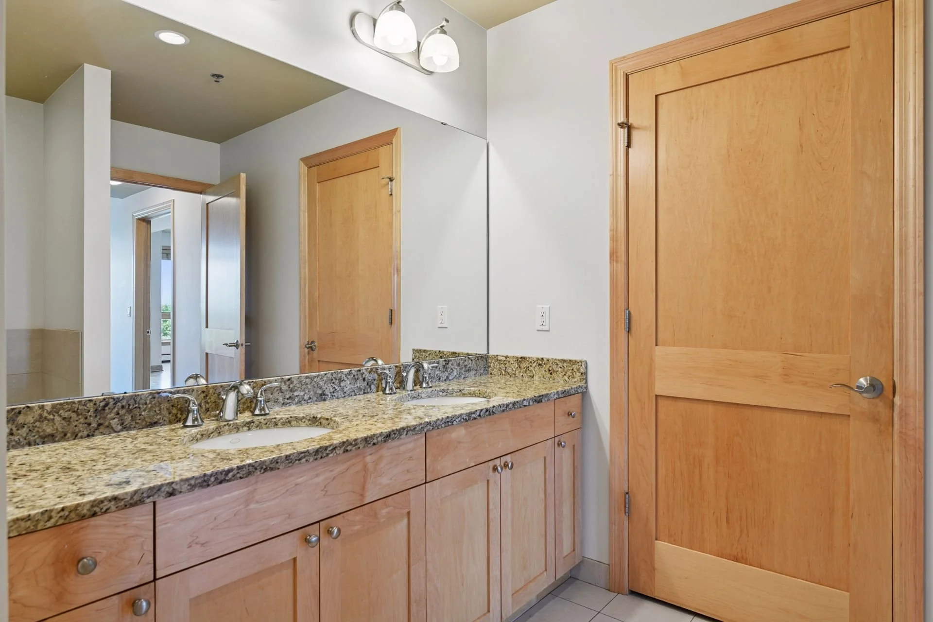 Bathroom with a granite countertop and dual sinks, large mirror, wooden cabinets, and a door, with a glimpse of a room with a balcony outside.