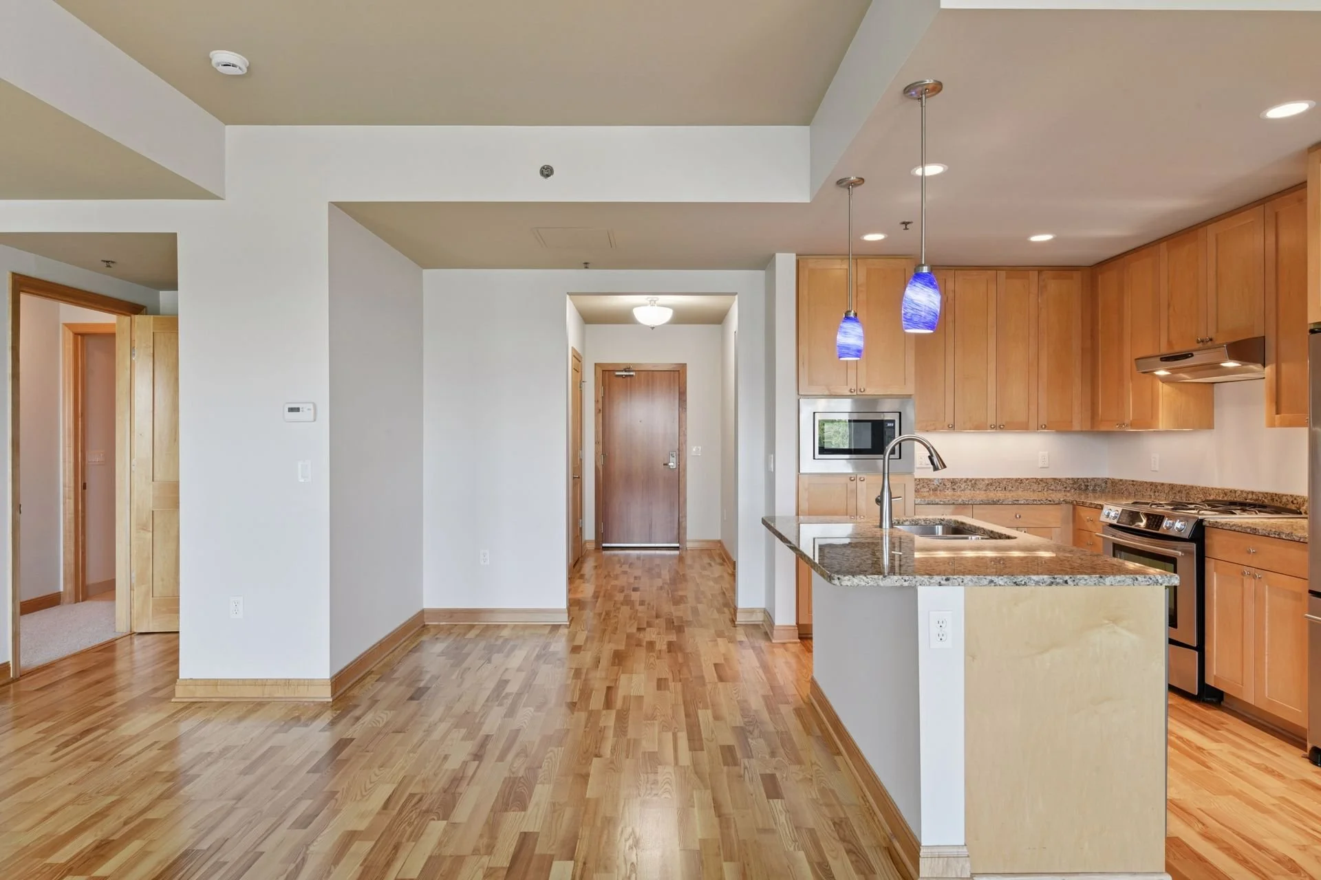 Empty kitchen and living space with hardwood floors, wooden cabinets, granite countertops, pendant lights, and an entrance door in the background.