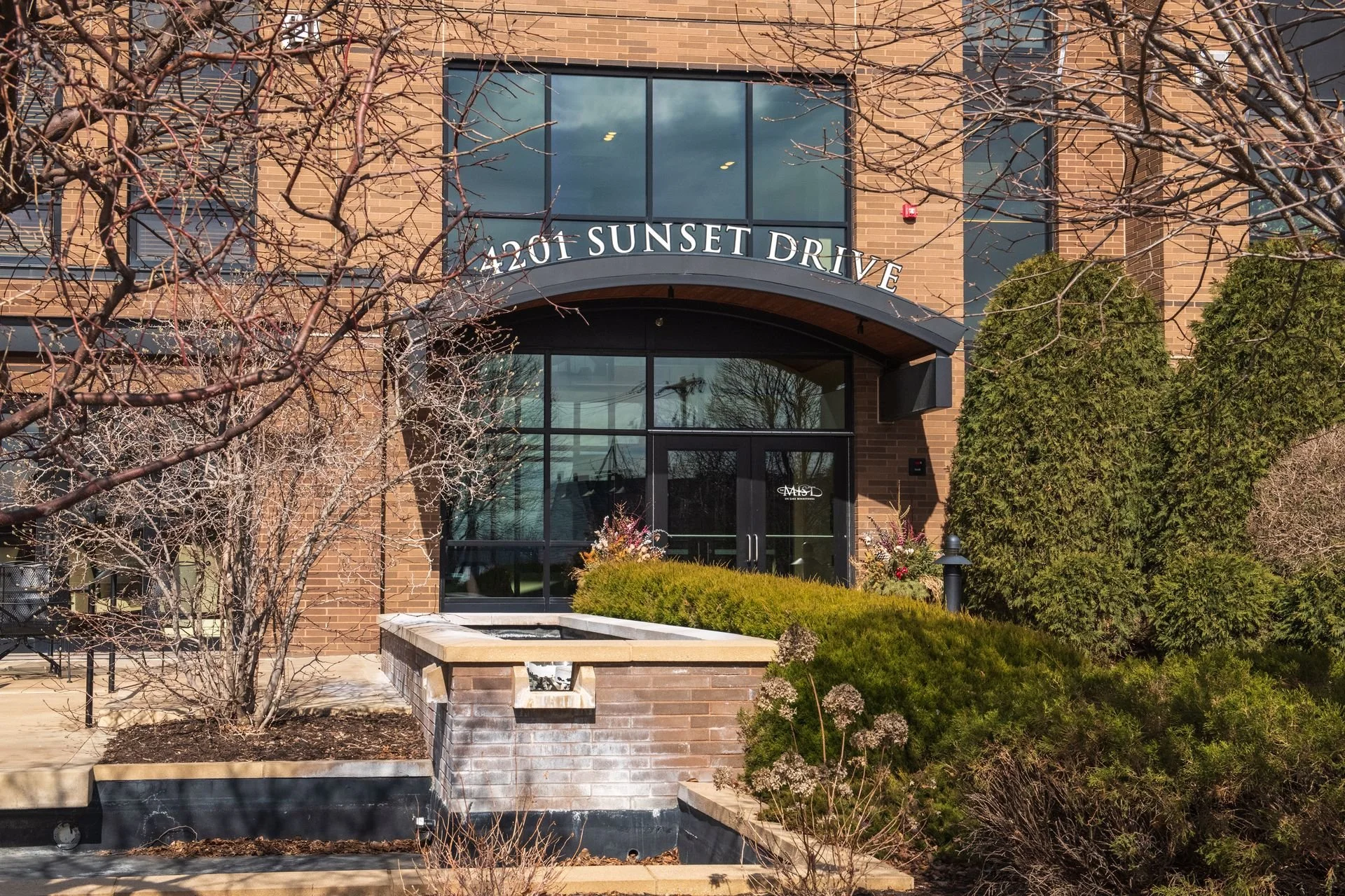 Entrance to a building with a sign that reads 4207 Sunset Drive, surrounded by trees and shrubs, with a flower bed near the door.