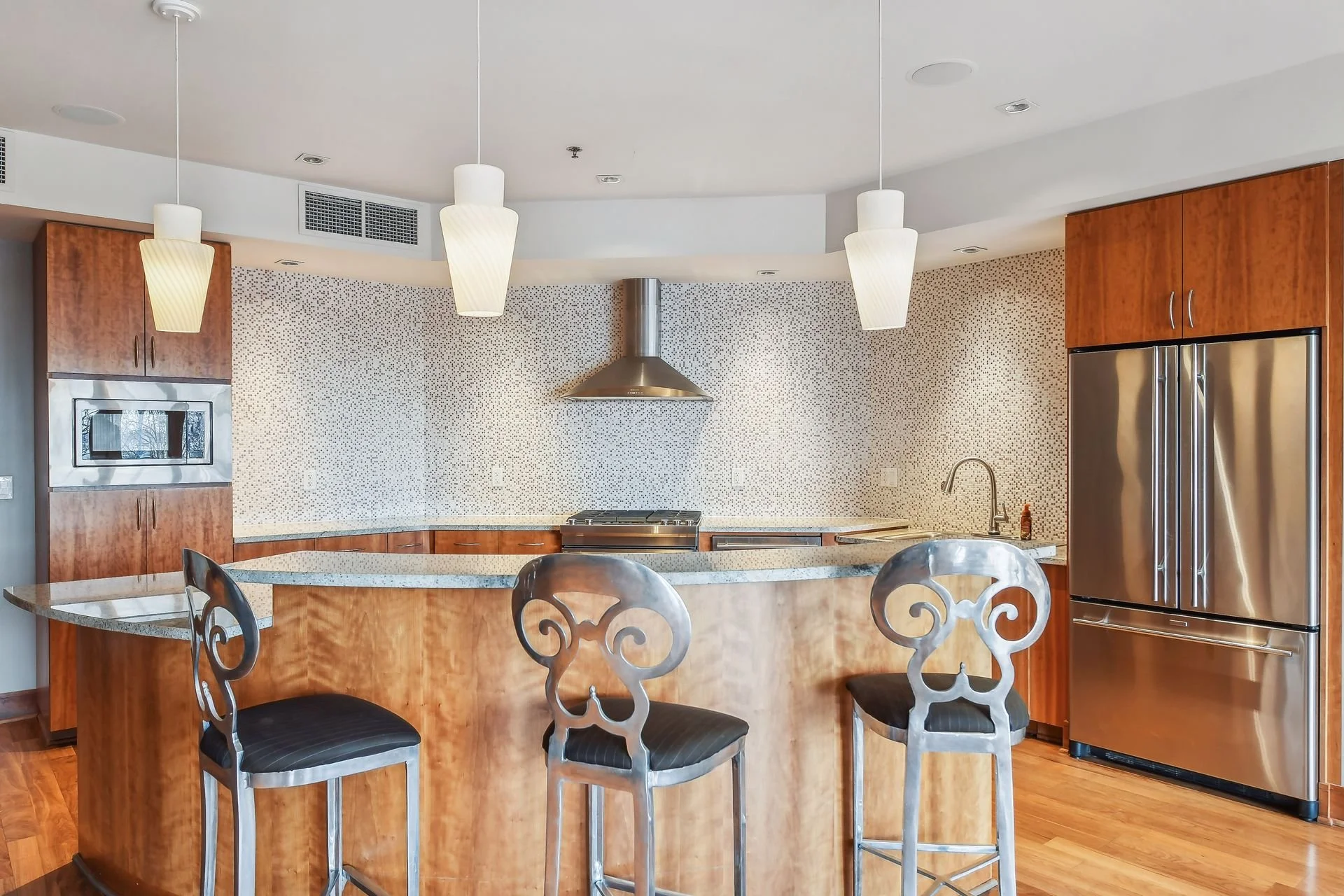 Community room's modern kitchen with wooden cabinets, stainless steel refrigerator and microwave, granite island with three decorative chairs, patterned backsplash, and pendant lighting.
