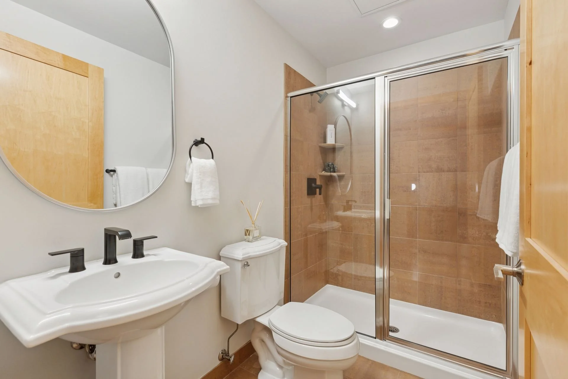 Bathroom with a white sink, black faucet, oval mirror, toilet, and a glass shower with earthy brown tiles.