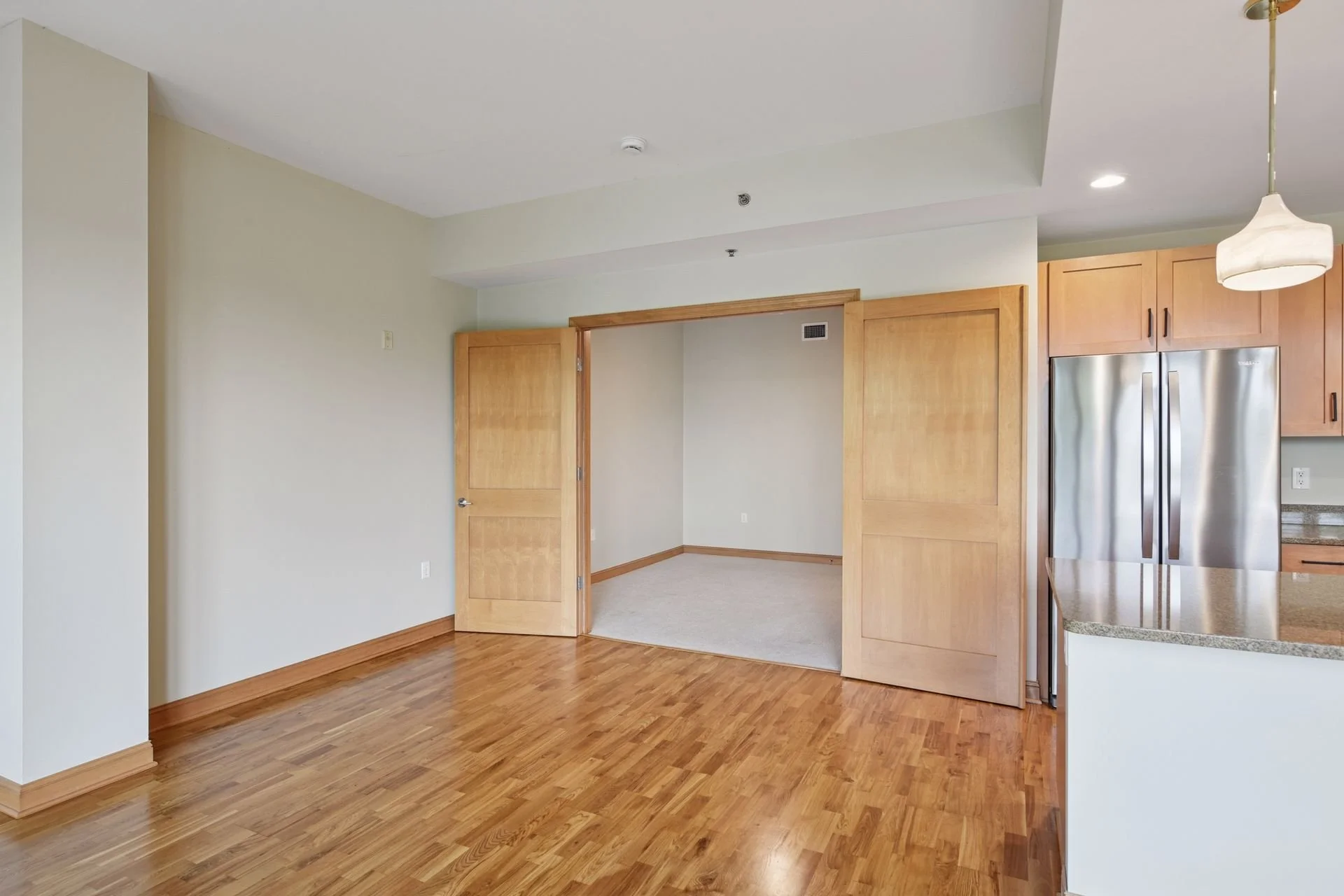 Empty living room with hardwood floors, an open doorway leading to another room, and kitchen cabinets with a refrigerator.