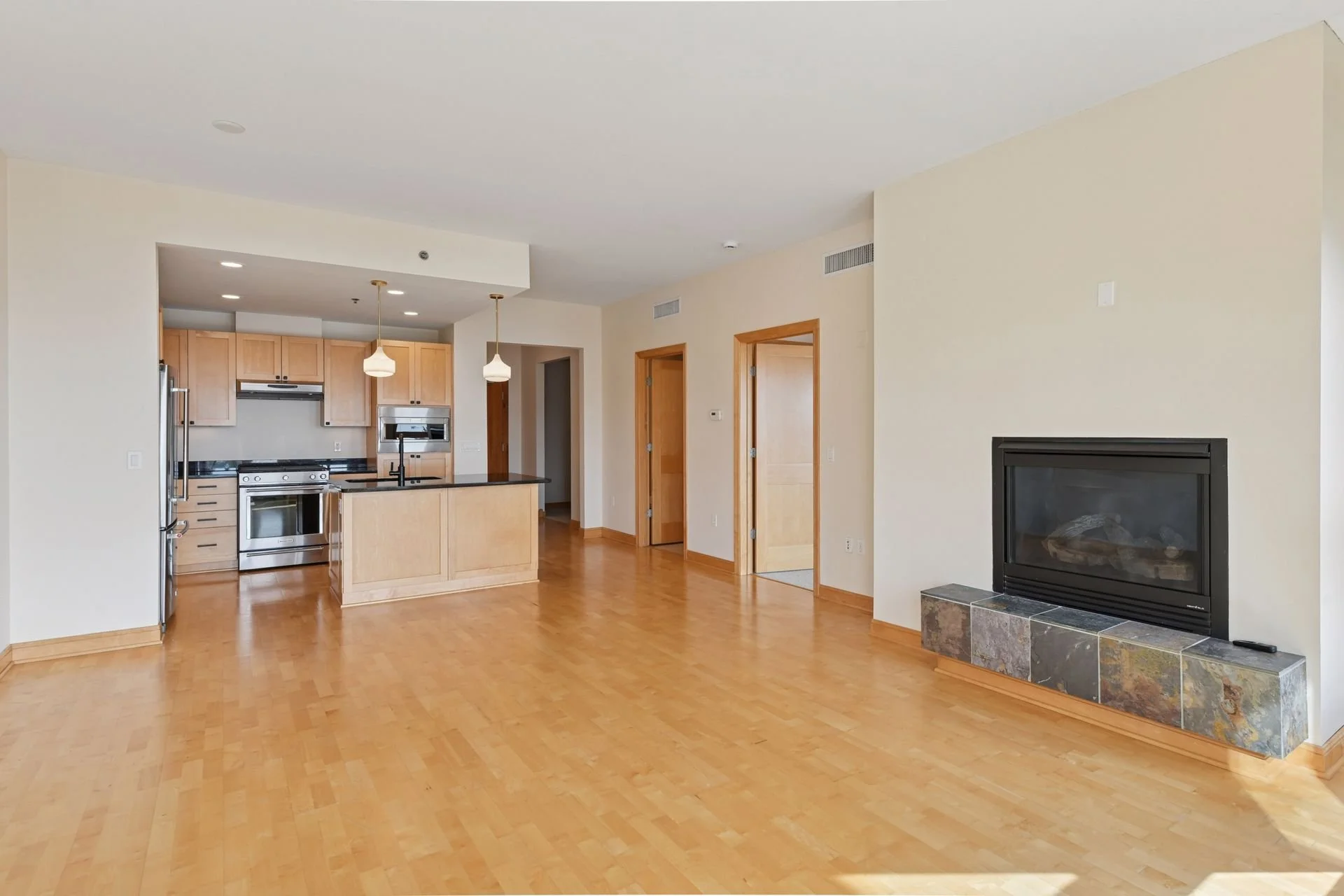 Empty living room with hardwood floors, white walls, a fireplace with a stone mantel, and an open kitchen with light wood cabinets, black countertops, and pendant lighting.