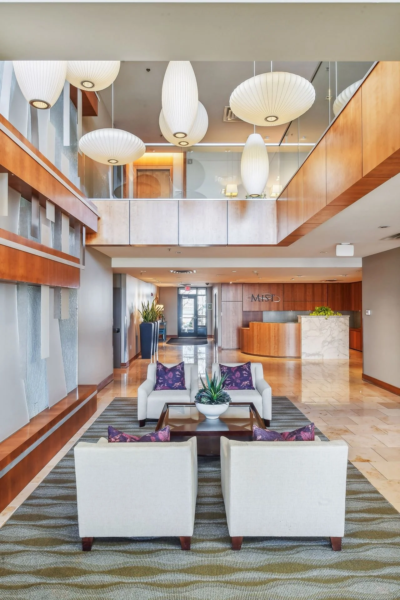 Lobby with white sofas, purple pillows, a glass table, large hanging white lanterns, wooden accents, and a reception desk in the background.