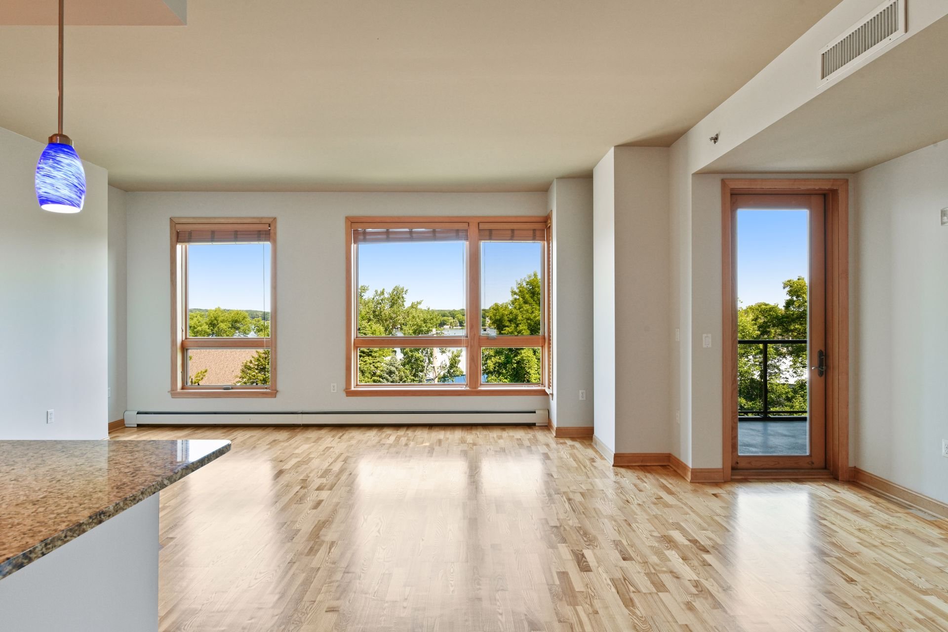 Living room with three large windows, a glass door leading to a balcony, wooden flooring, and a blue pendant light