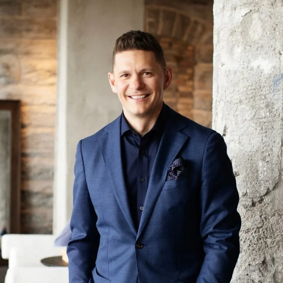 A smiling man wearing a dark blue suit with a pocket square, standing indoors against a textured concrete wall and wooden wall background.