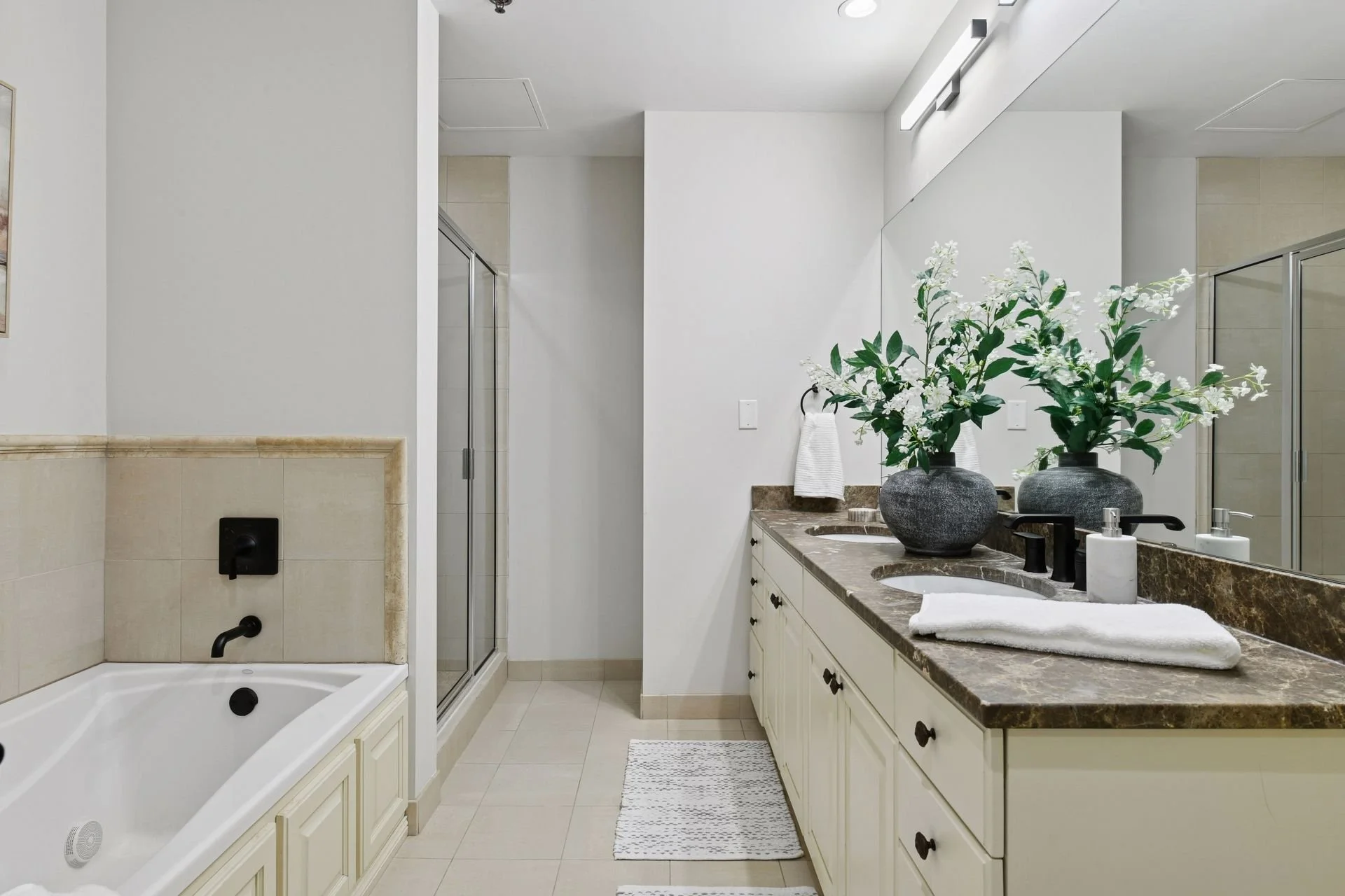 Modern bathroom with white walls, beige tile flooring, a bathtub on the left, a glass shower in the center, and a long vanity on the right with a brown marble countertop, and a mirror.