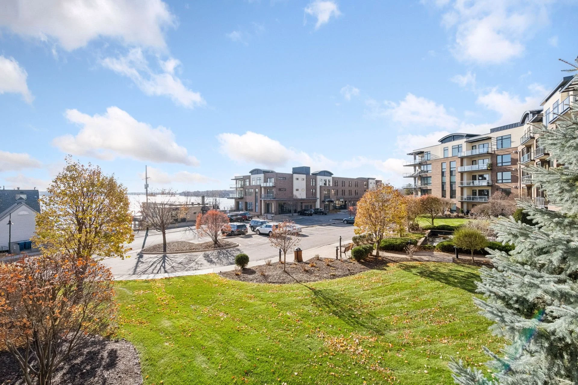 View of a cityscape with modern apartment buildings, parking lot with cars, and a landscaped area with trees and grass on a clear day with blue sky and clouds.