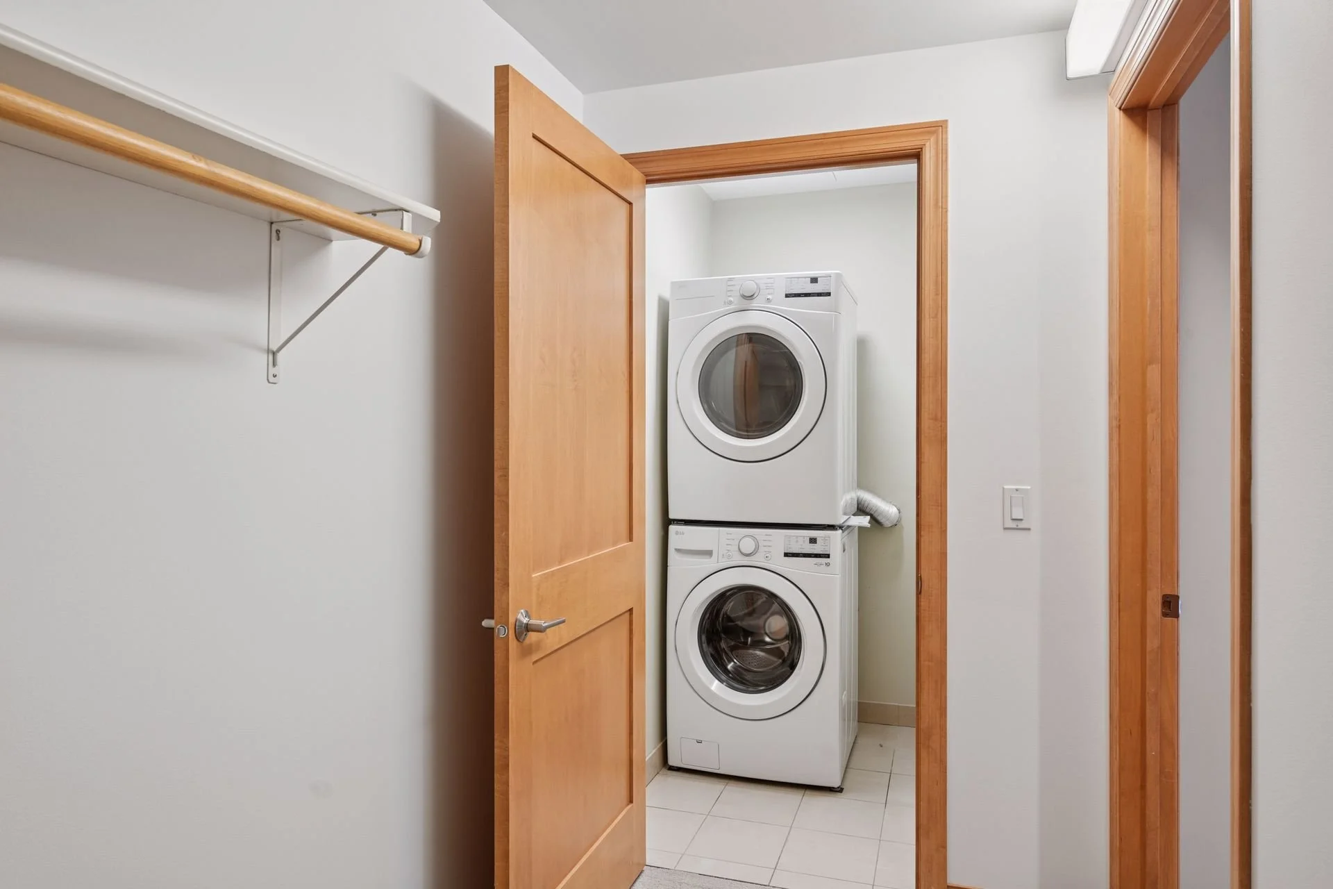 A laundry room with a stacked washer and dryer behind a partially open wooden door, with a white wall and a wooden closet rod on the left.