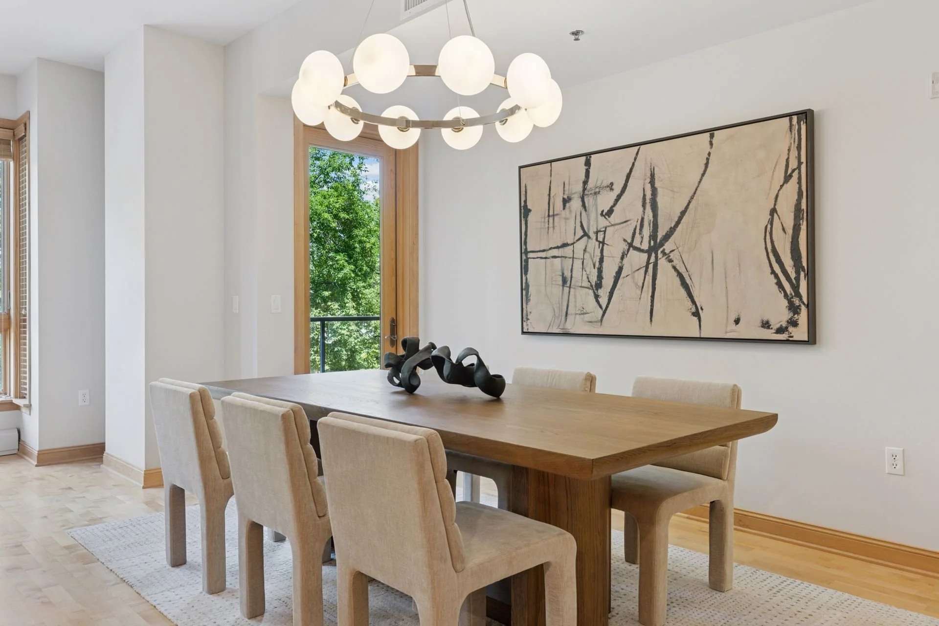 Modern dining room with a large circular chandelier with globes, and a door leading to an outdoor balcony with green foliage visible.
