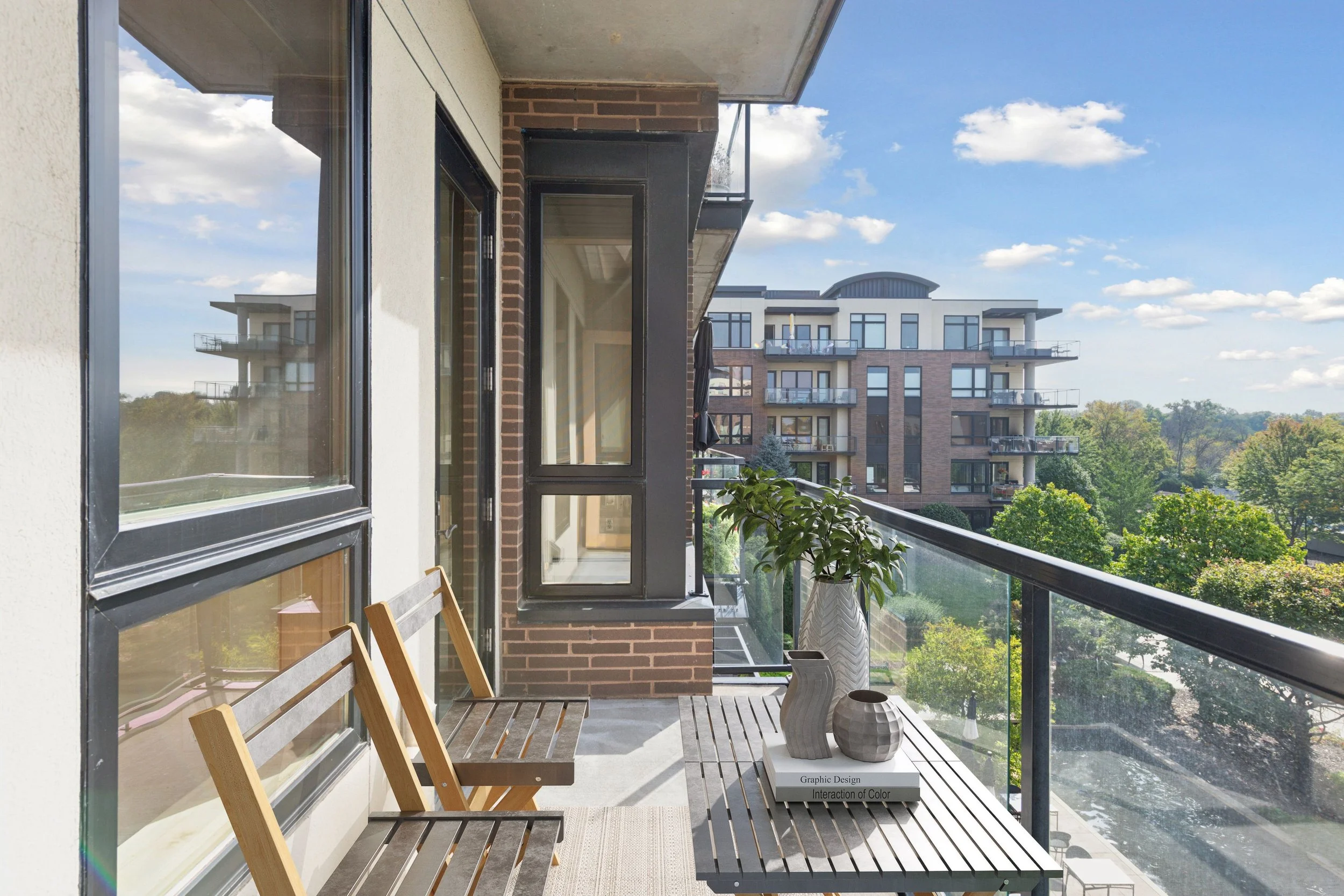 Balcony with wooden bench, potted plants, decorative vases, and a coffee table, overlooking a residential area with trees and another apartment building in the distance.