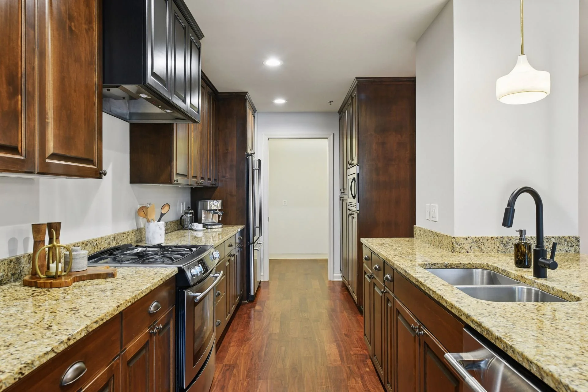 A modern kitchen with wooden cabinets, granite countertops, and stainless steel appliances. There is a sink with a black faucet on the right and a stove with an oven on the left. The kitchen has hardwood flooring and is well-lit with ceiling lights a