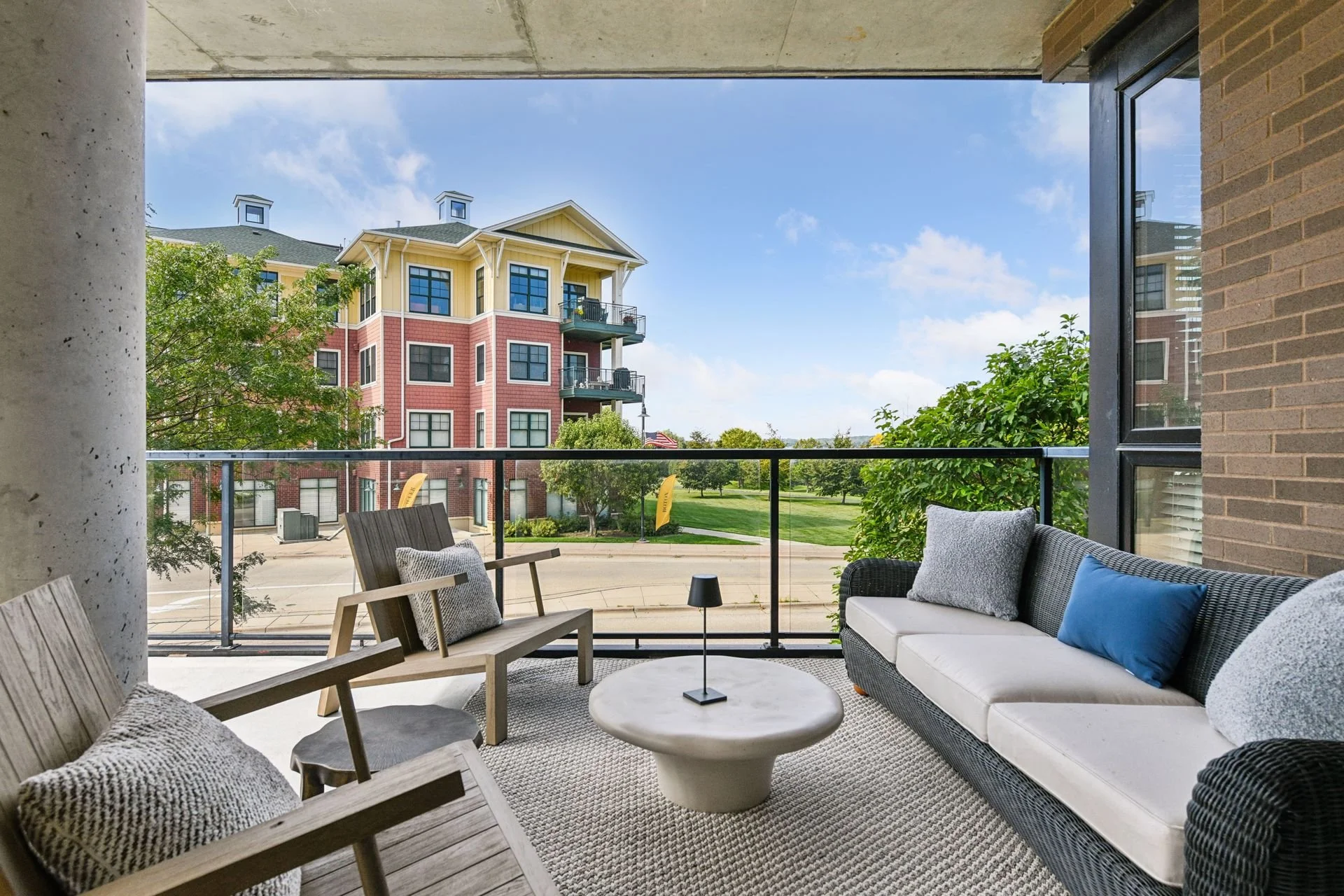 Outdoor balcony overlooking a street and colorful apartment building with balconies, trees, and a green lawn, under a blue sky with scattered clouds.