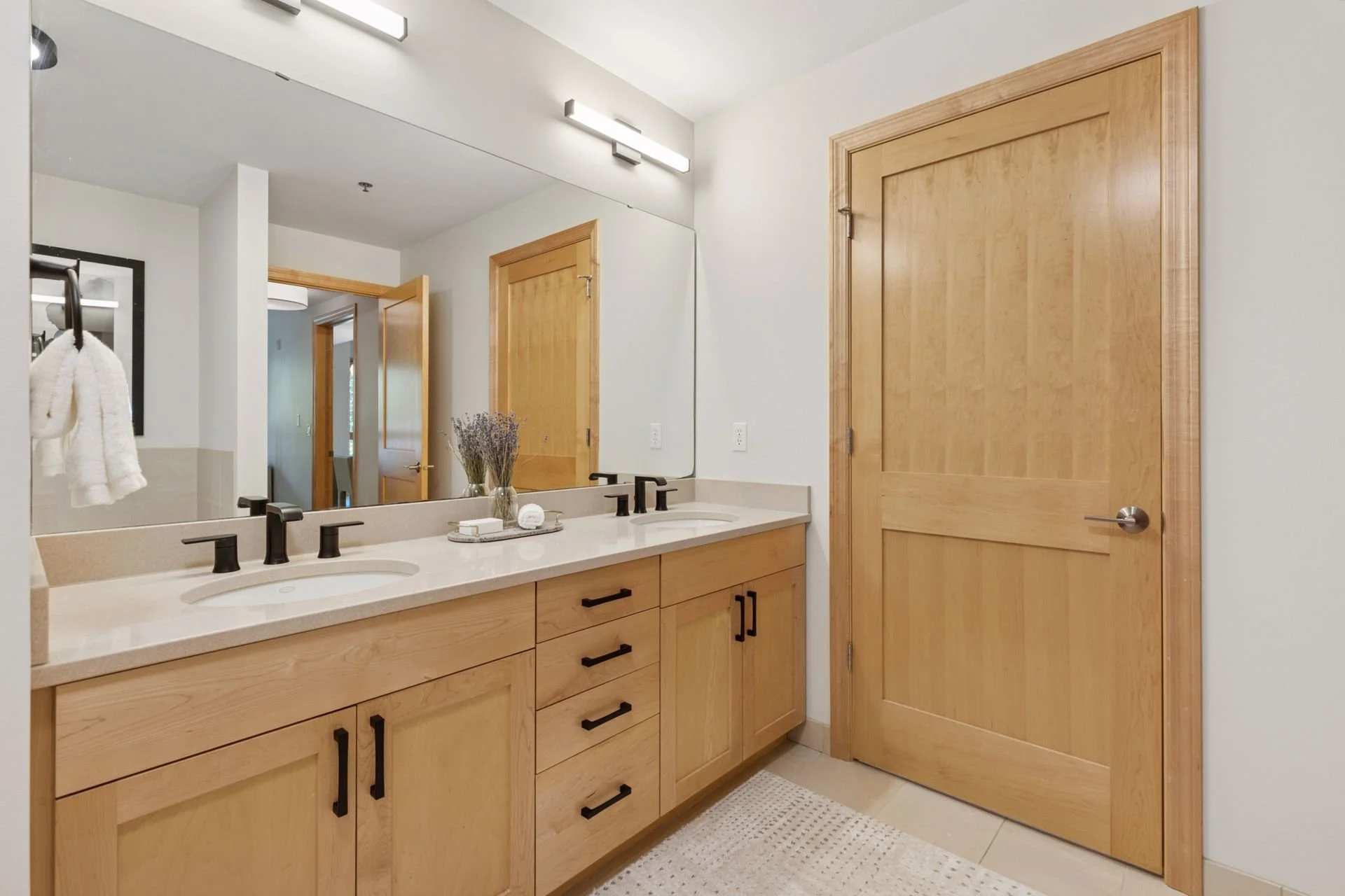 Bathroom with double vanity sink, large mirror, light wood cabinetry, black fixtures, light-colored countertop, wooden door, and beige tile flooring.