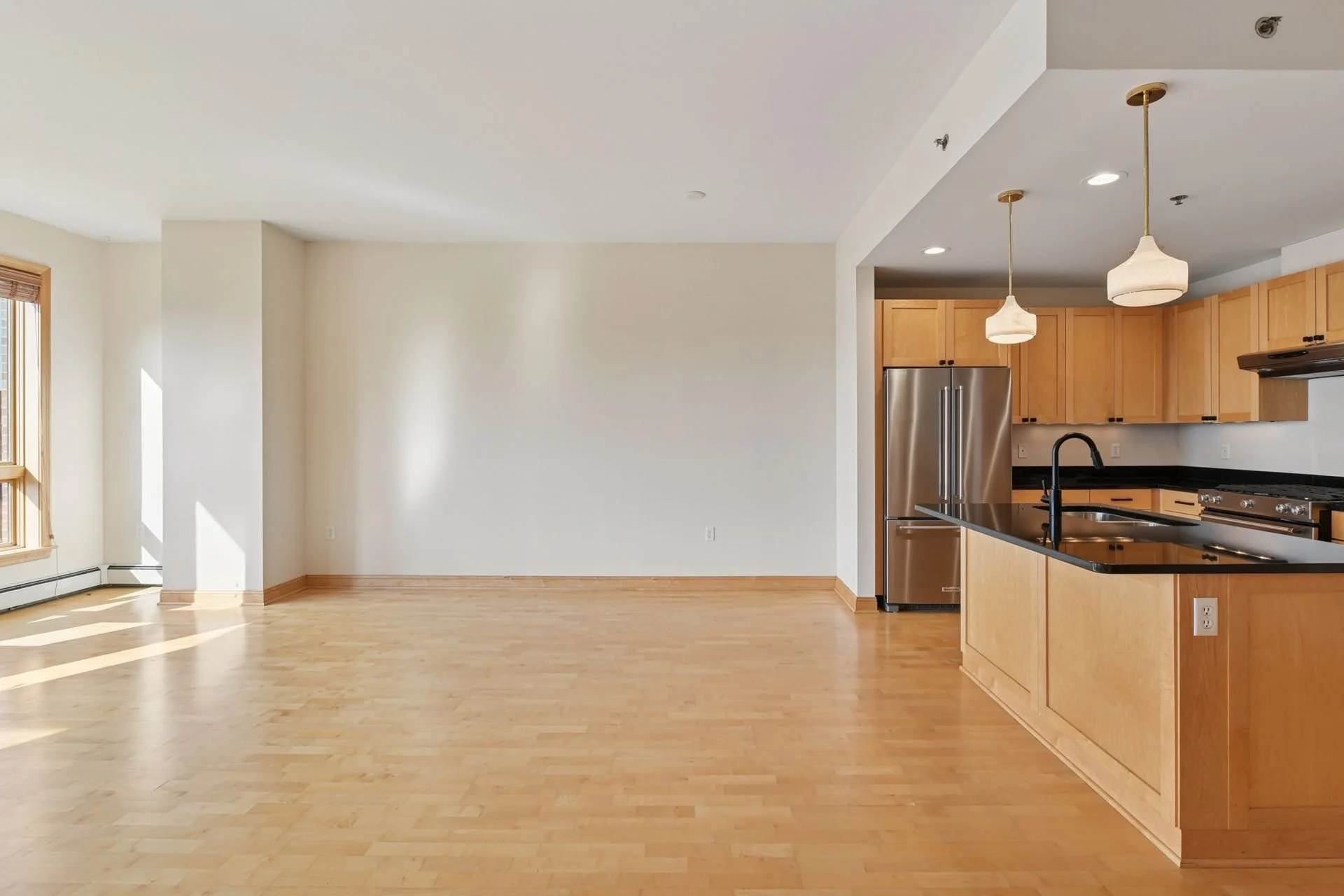 Empty living room with hardwood floors and a view of the kitchen with wooden cabinets, stainless steel refrigerator, black countertop, and pendant lights.
