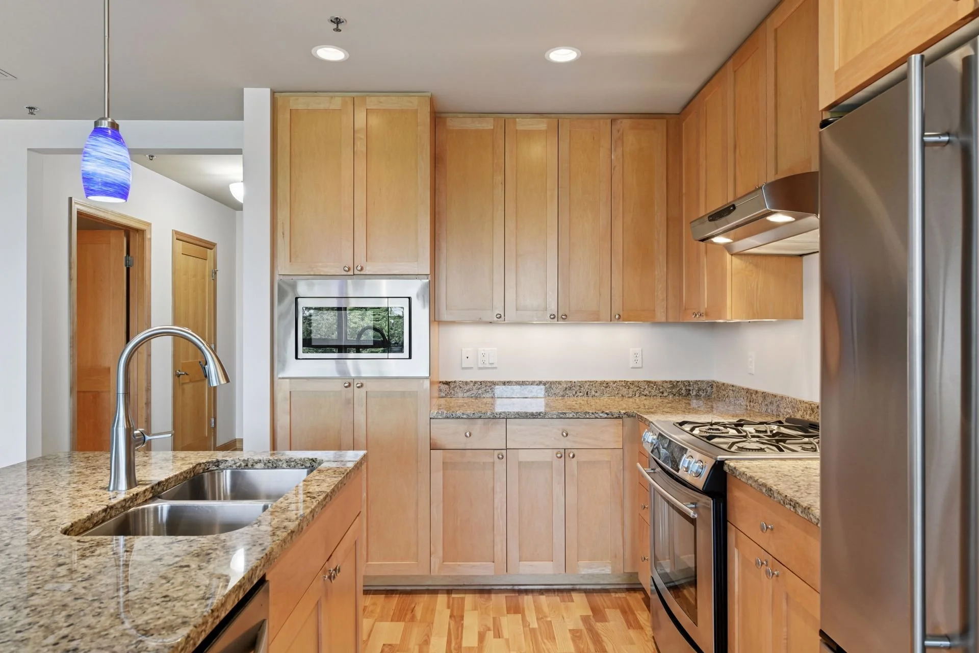 Modern kitchen with light wooden cabinets, granite countertops, stainless steel appliances including a refrigerator and stove, a double sink, and ceiling lighting.