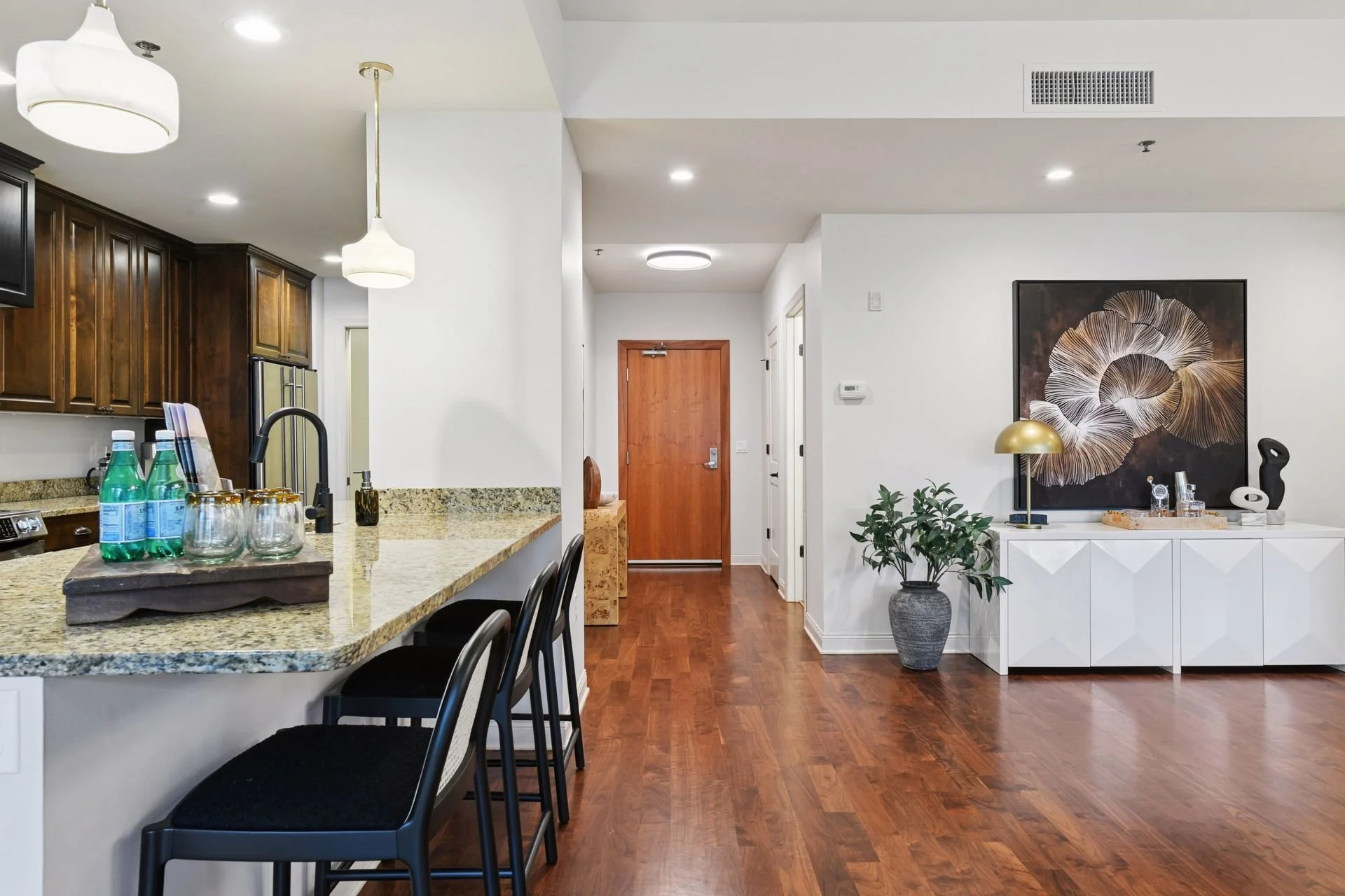 Interior view of a modern apartment with a kitchen and living area. The kitchen has a granite countertop, dark wooden cabinets, and hanging pendant lights. The living area includes hardwood floors.