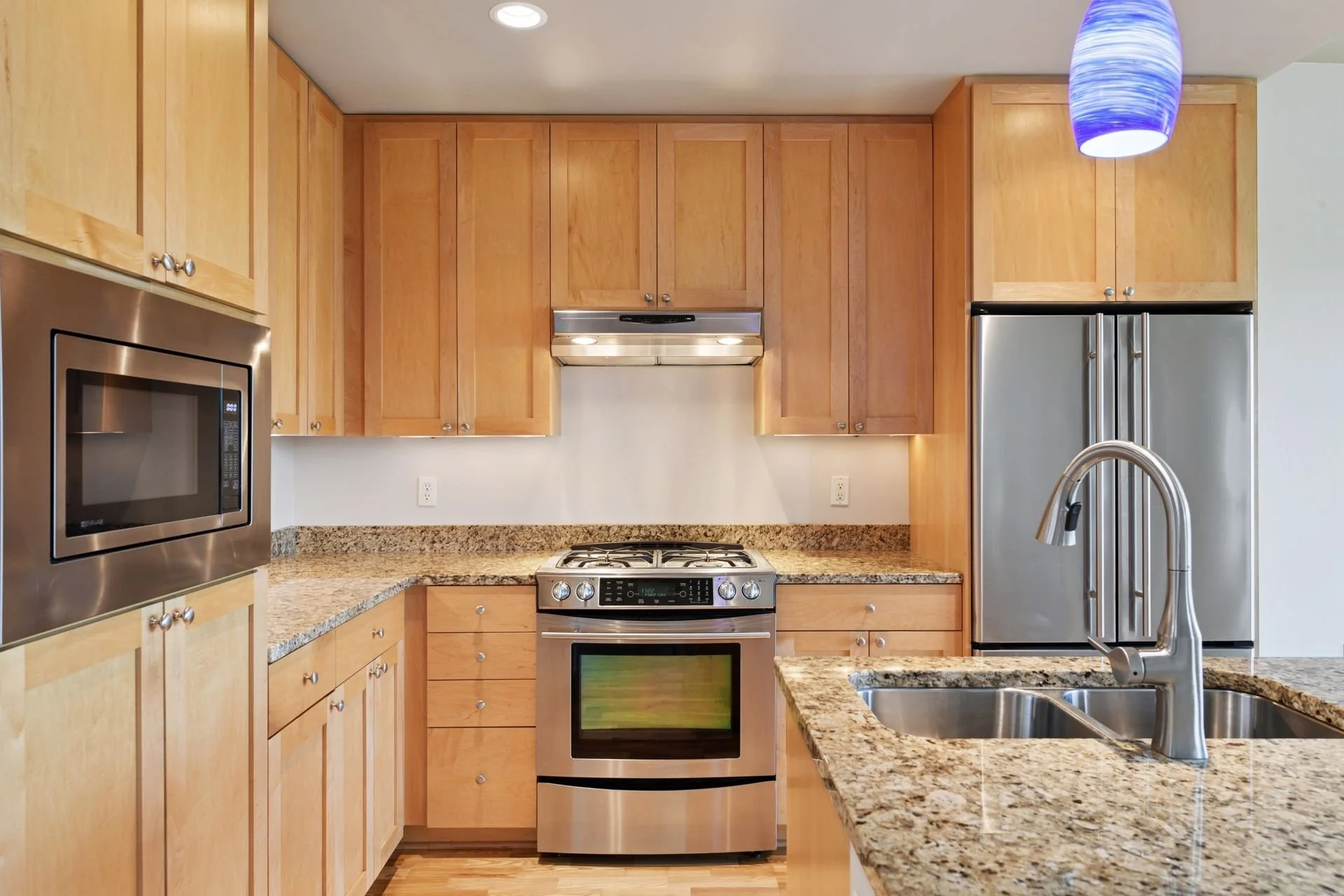 Kitchen with wooden cabinets, granite countertops, stainless steel appliances including a refrigerator, microwave, and oven, with a kitchen sink and a blue hanging light fixture.