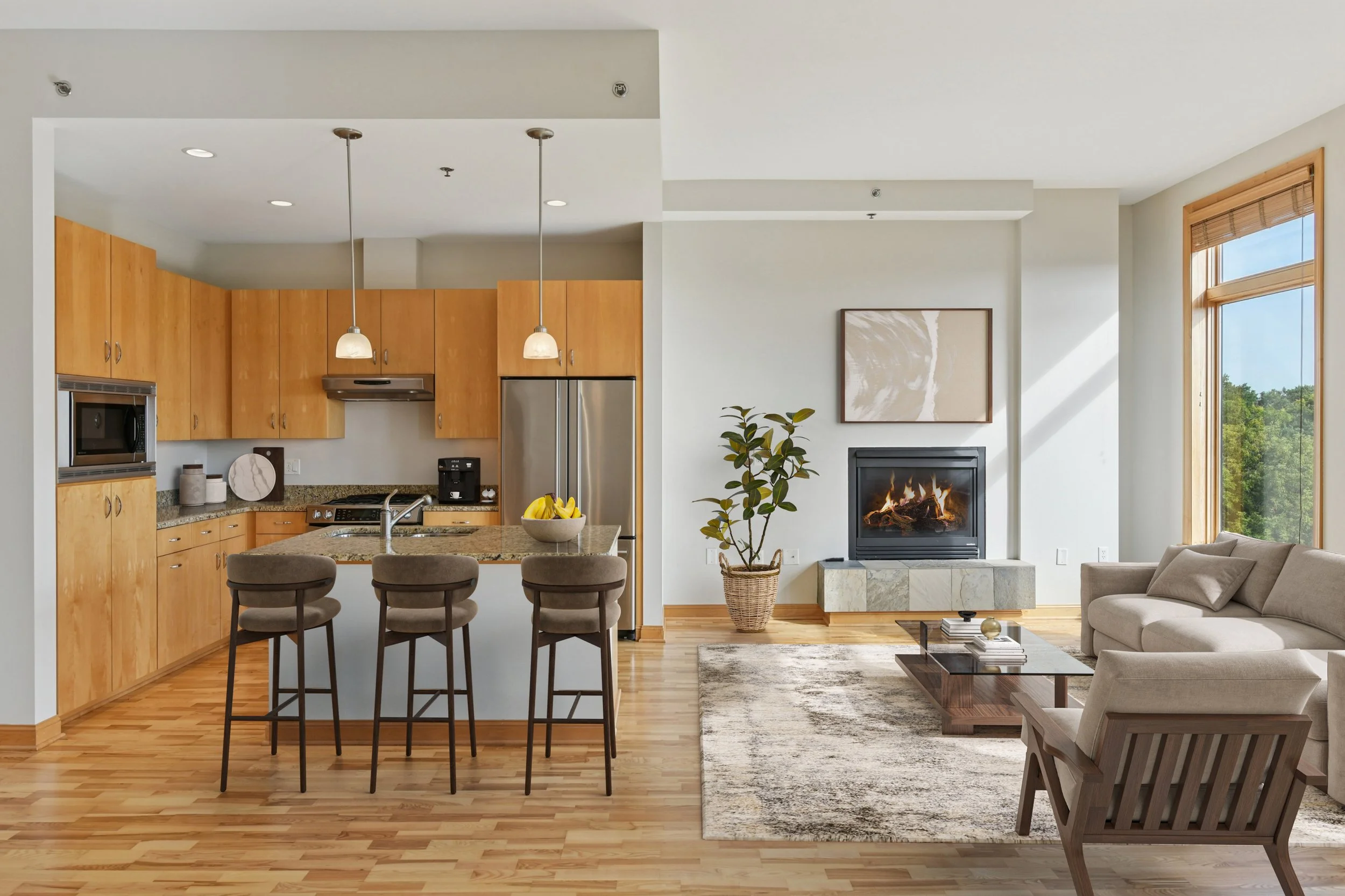 Open-concept living room and kitchen with wooden flooring, fireplace, and large windows showing green trees outside.