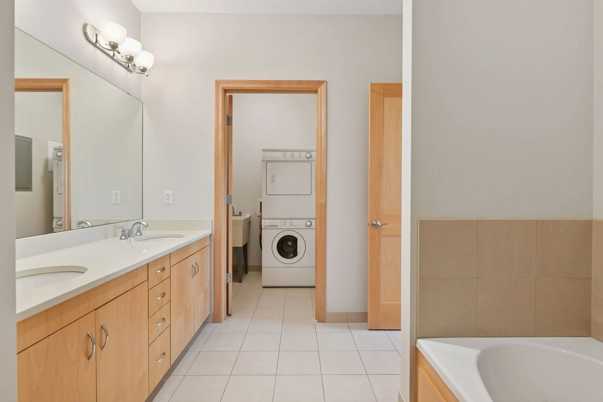 Bathroom with double vanity sink, large mirror, overhead lighting, washer and dryer in laundry area, part of a bathtub, tiled walls and floor, and wooden doors and cabinets.