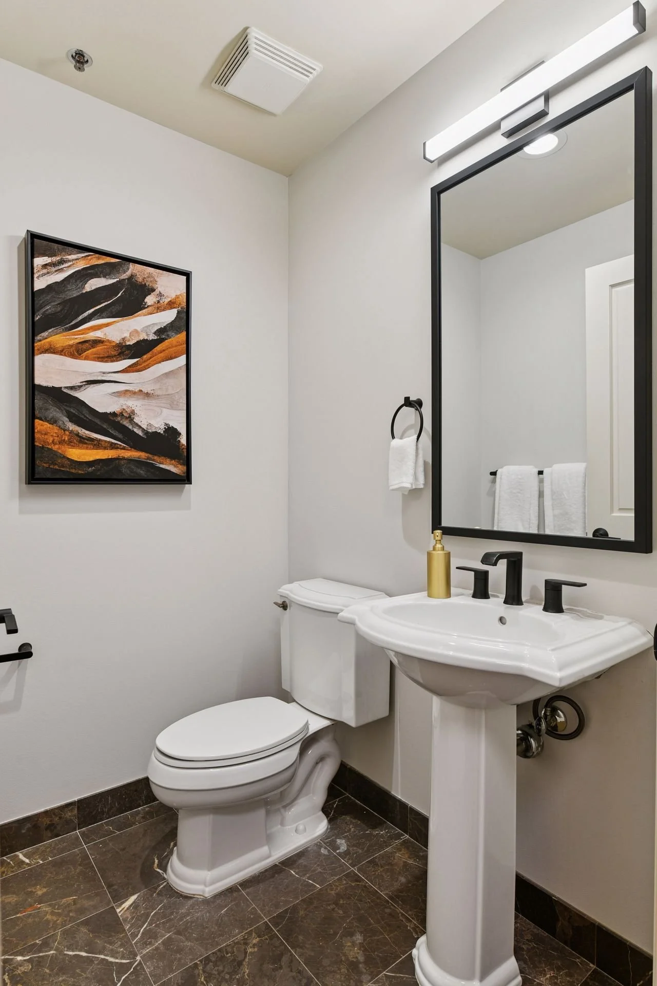 A bathroom with a toilet, a white pedestal sink with black hardware, a large mirror, and a modern strip light above the mirror. The floor is dark marble.