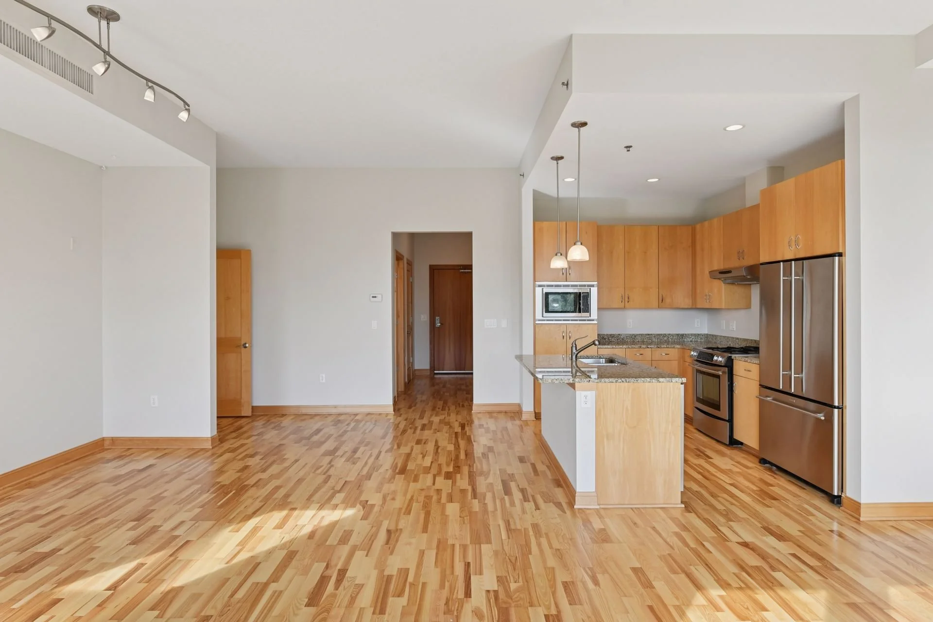 Empty apartment with hardwood floors, white walls, and a kitchen with wooden cabinets, stainless steel appliances, and granite countertops.