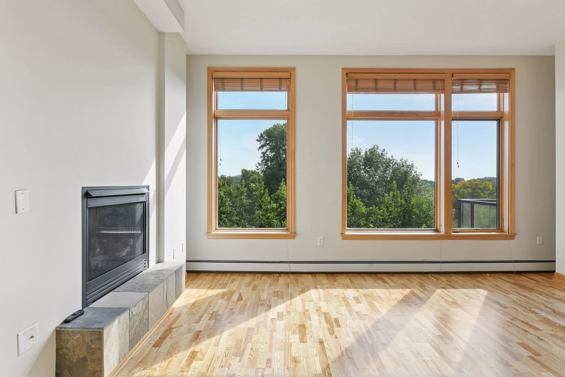 Empty living room with large windows showing a view of trees outdoors, a fireplace on the left, light hardwood floors, and white walls.