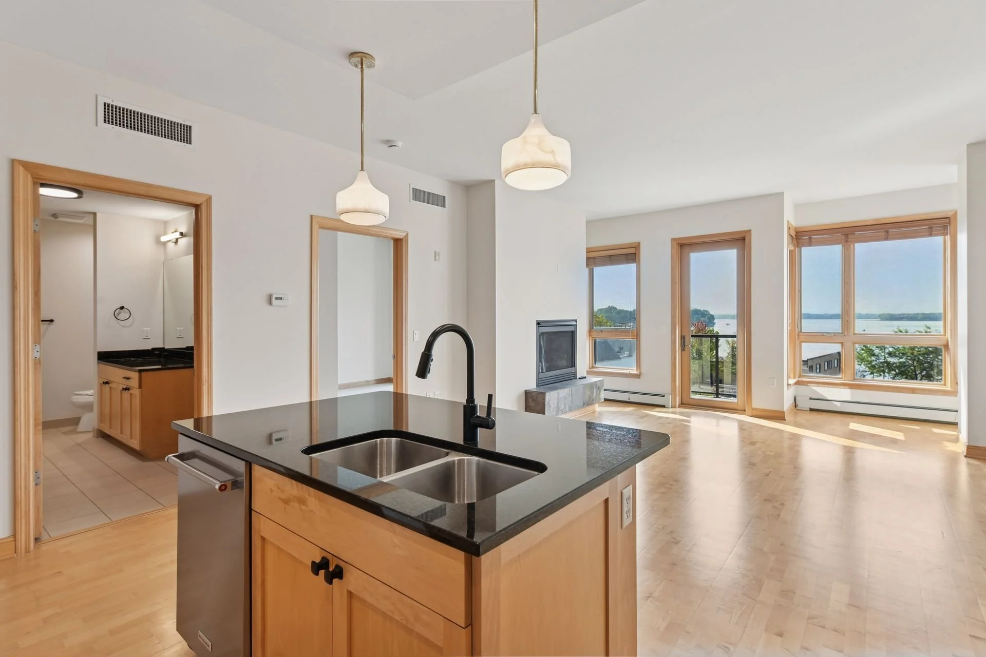 Bright living room with large windows overlooking water, open kitchen with island, black sink, and two hanging pendant lights.