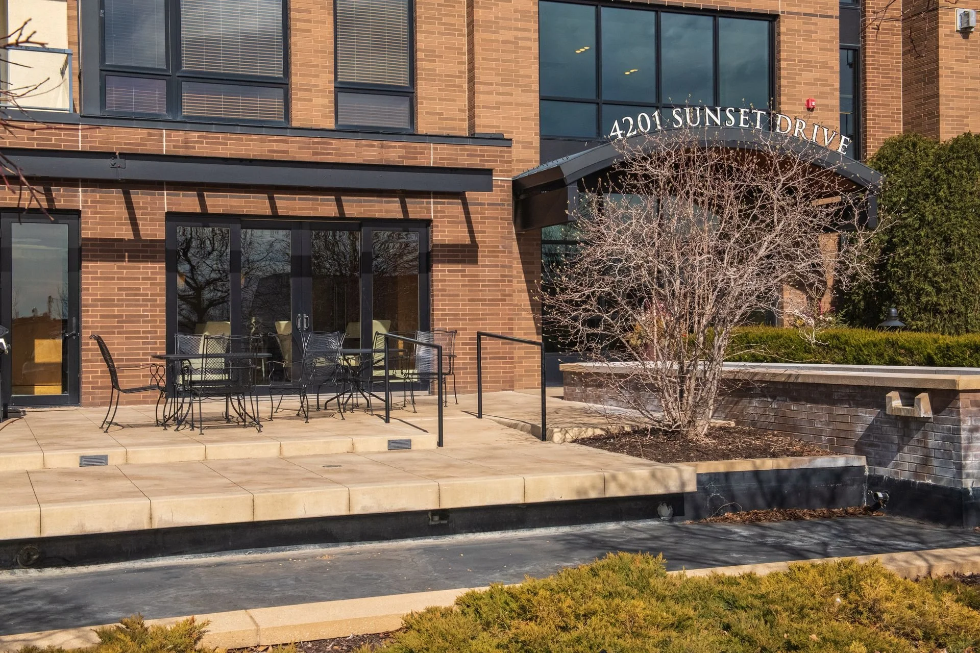 Exterior of a brick building with large glass windows, a small patio with chairs and tables, a leafless tree, and a sign reading '4201 Sunset Drive'