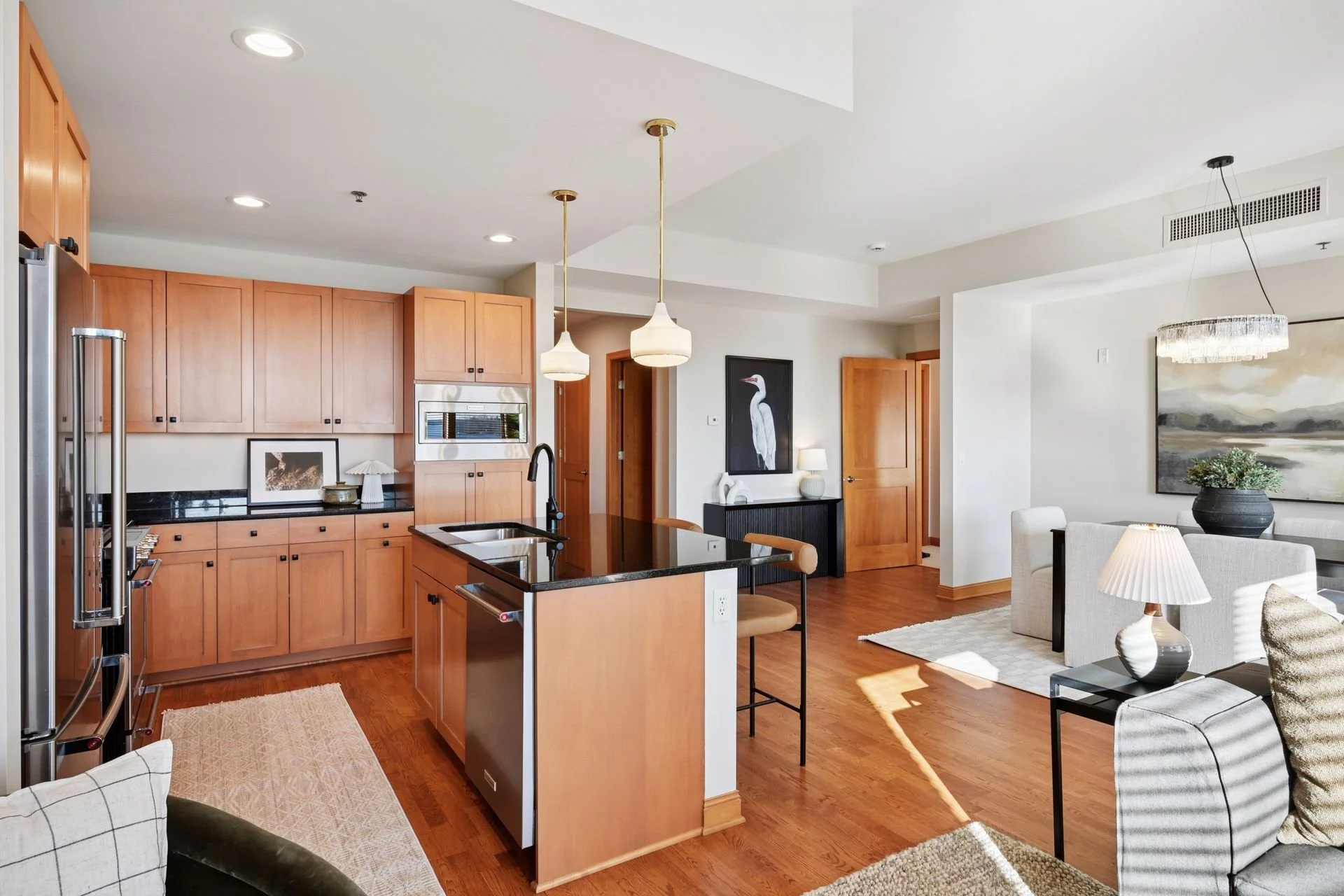 L-shaped open-concept living room and kitchen with wooden cabinetry, black countertops, hardwood floors, and white walls. The kitchen features a central island with a sinkand modern pendant lighting.