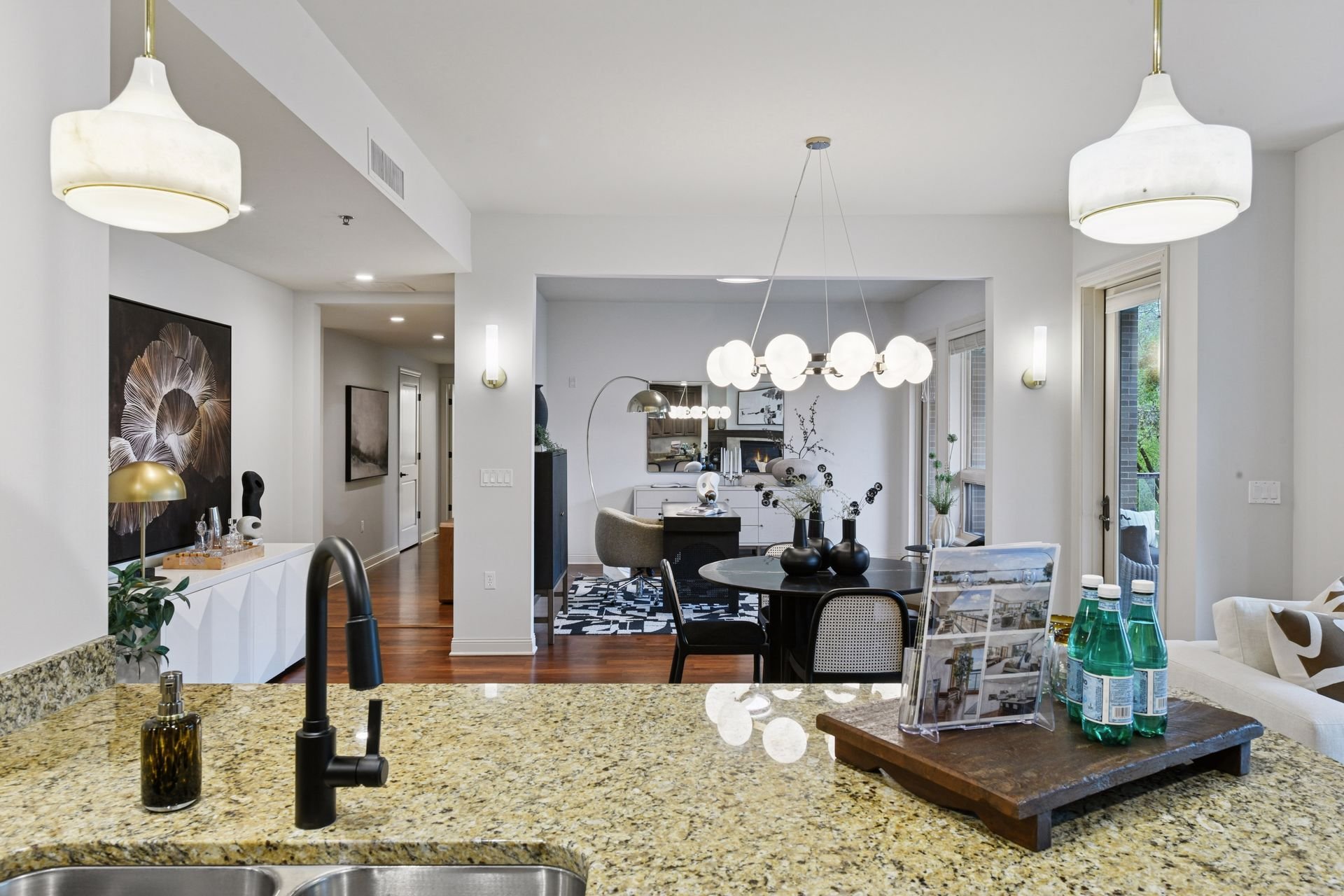 Modern kitchen and dining area with white walls and multiple hanging light fixtures, a granite countertop with a sink, and a living space.