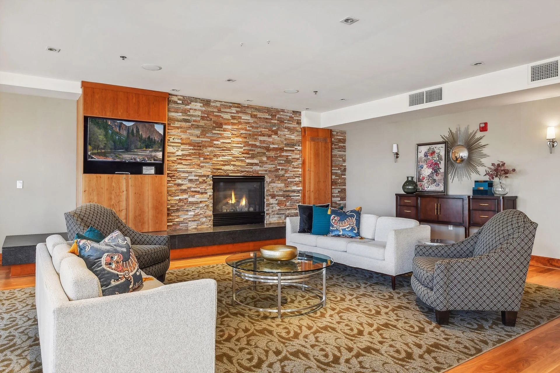 Community room with stone fireplace, sectional and armchairs, decorative pillows, wall-mounted TV, sideboard with vases and artwork, carpet, and wall sconces.