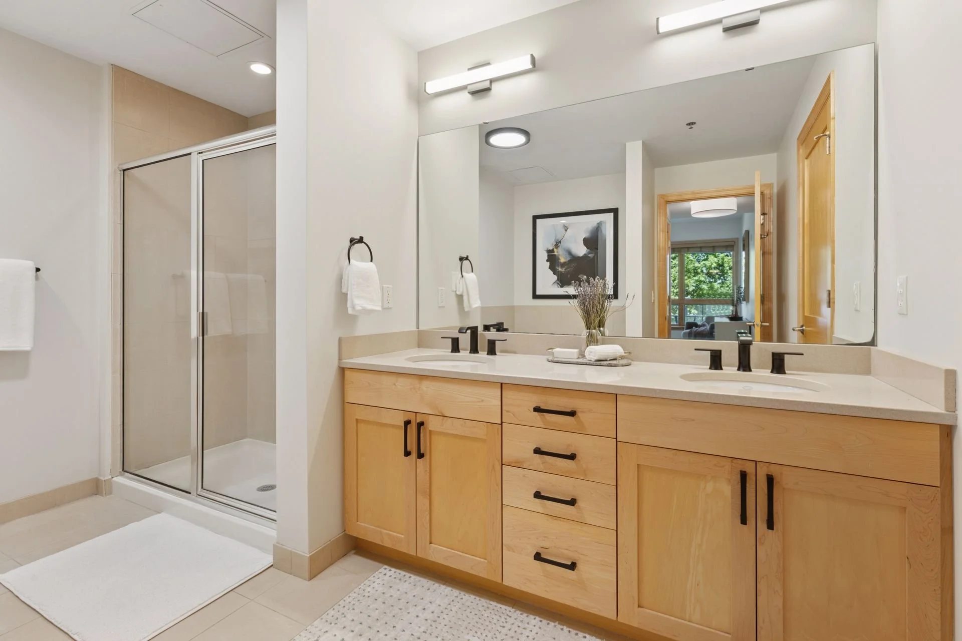 Bathroom with double vanity, large mirror, black faucets, and wooden cabinets, a shower with glass door, and a view into a living area with a sliding door and outdoor greenery.