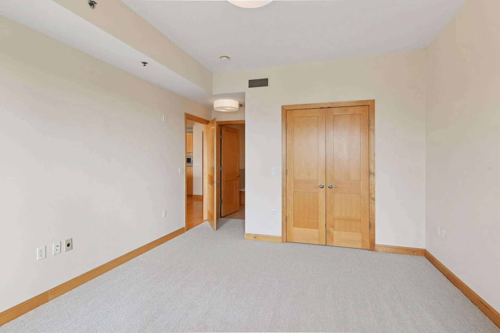 An empty room with beige walls, a beige carpet, and wooden trim. There are two wooden doors, one open and one closed. An open door reveals a hallway and kitchen area.