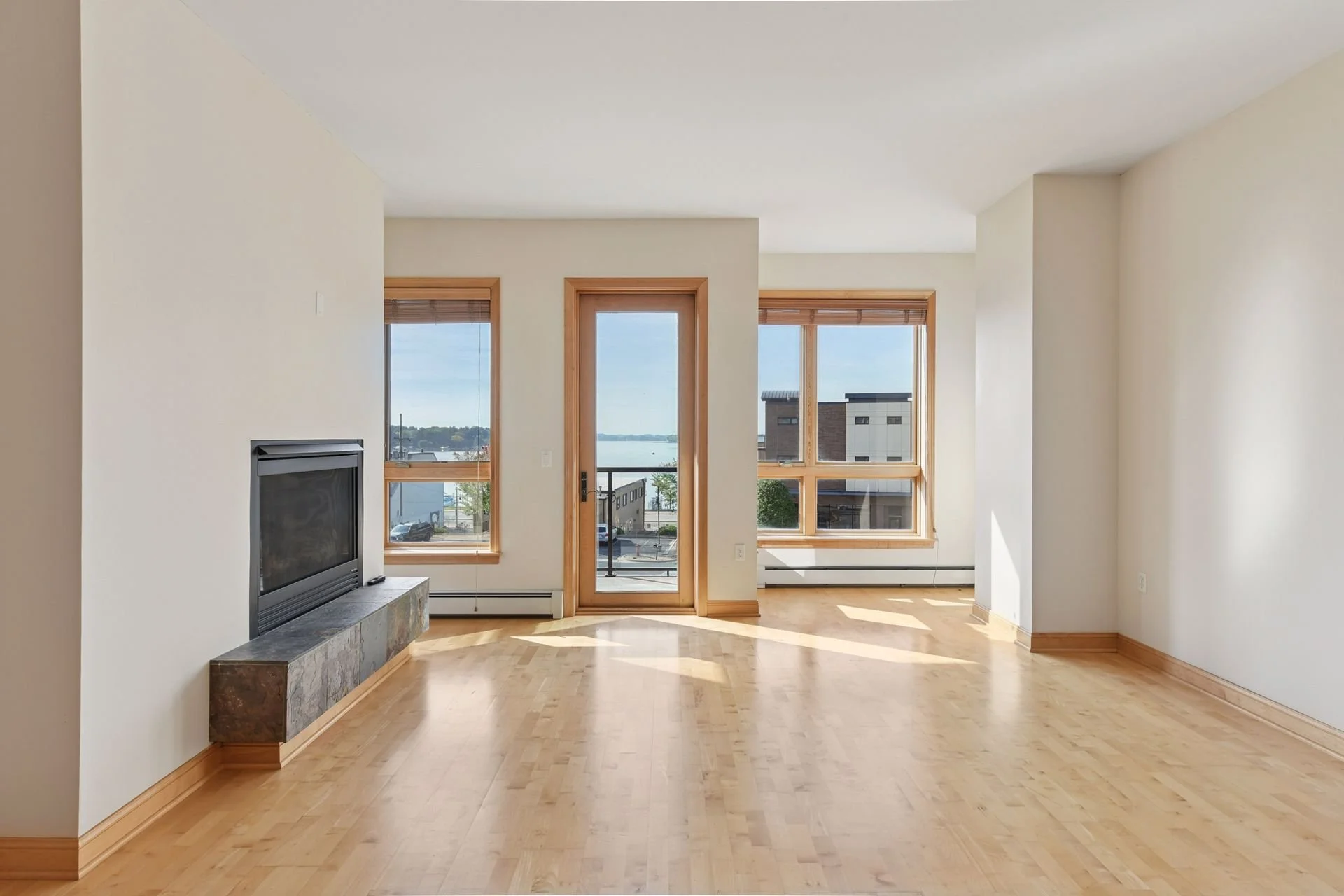 Empty living room with hardwood floors, large windows, a glass door leading to a balcony, and a fireplace with a tiled hearth.