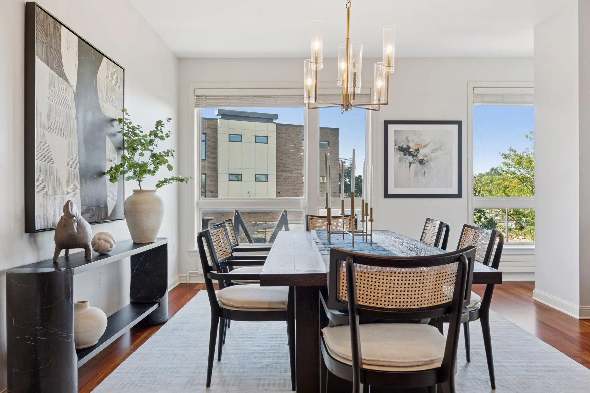 Modern dining room with a gold and glass chandelier, and large windows with views of trees and neighboring buildings.