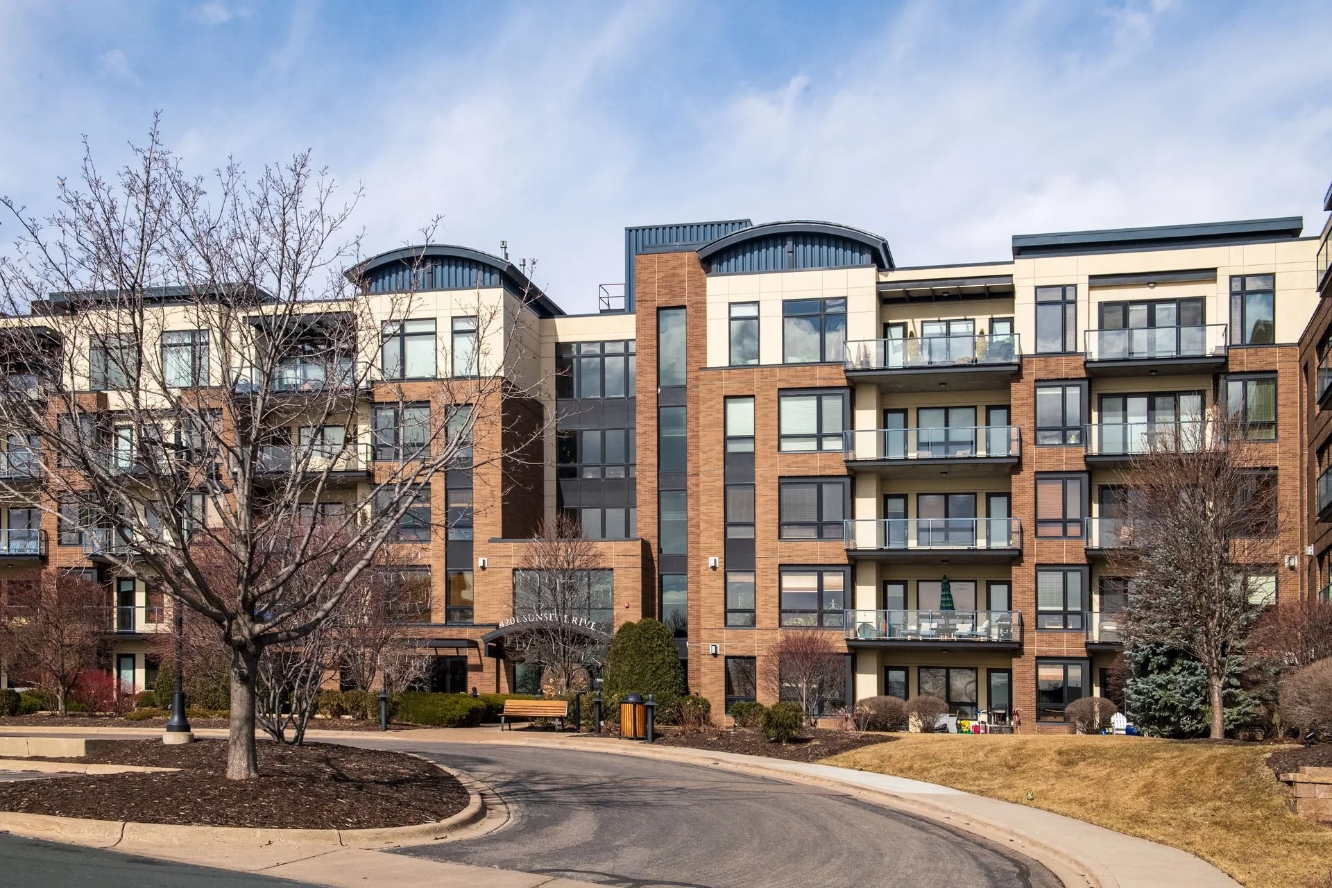 A modern multi-story apartment building with brick and beige exterior, multiple balconies, and large windows. There are landscaped grounds in front of the building under a partly cloudy sky.