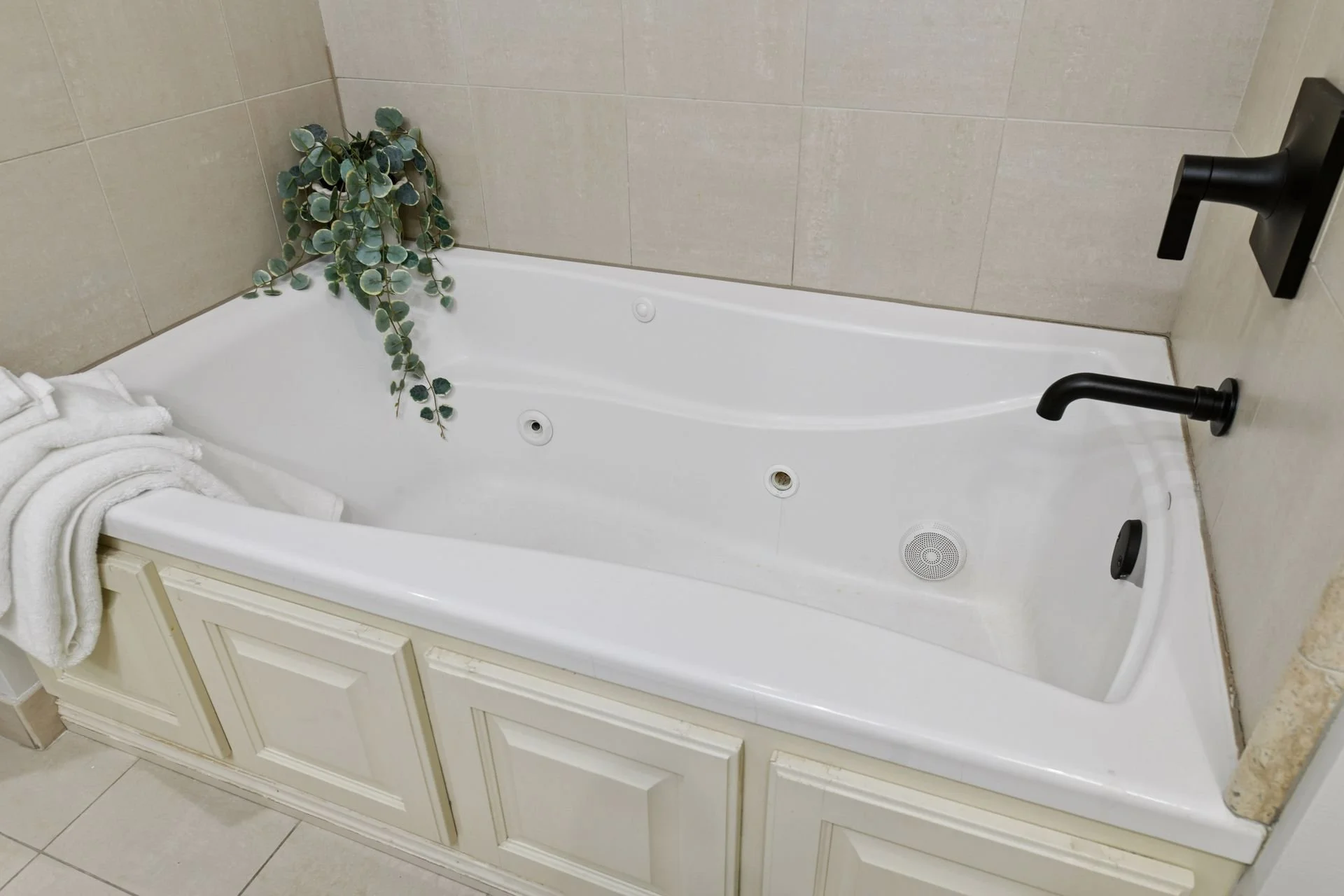 Empty white bathtub with black fixtures, a towel, and a green plant in a bathroom with beige tiled walls.