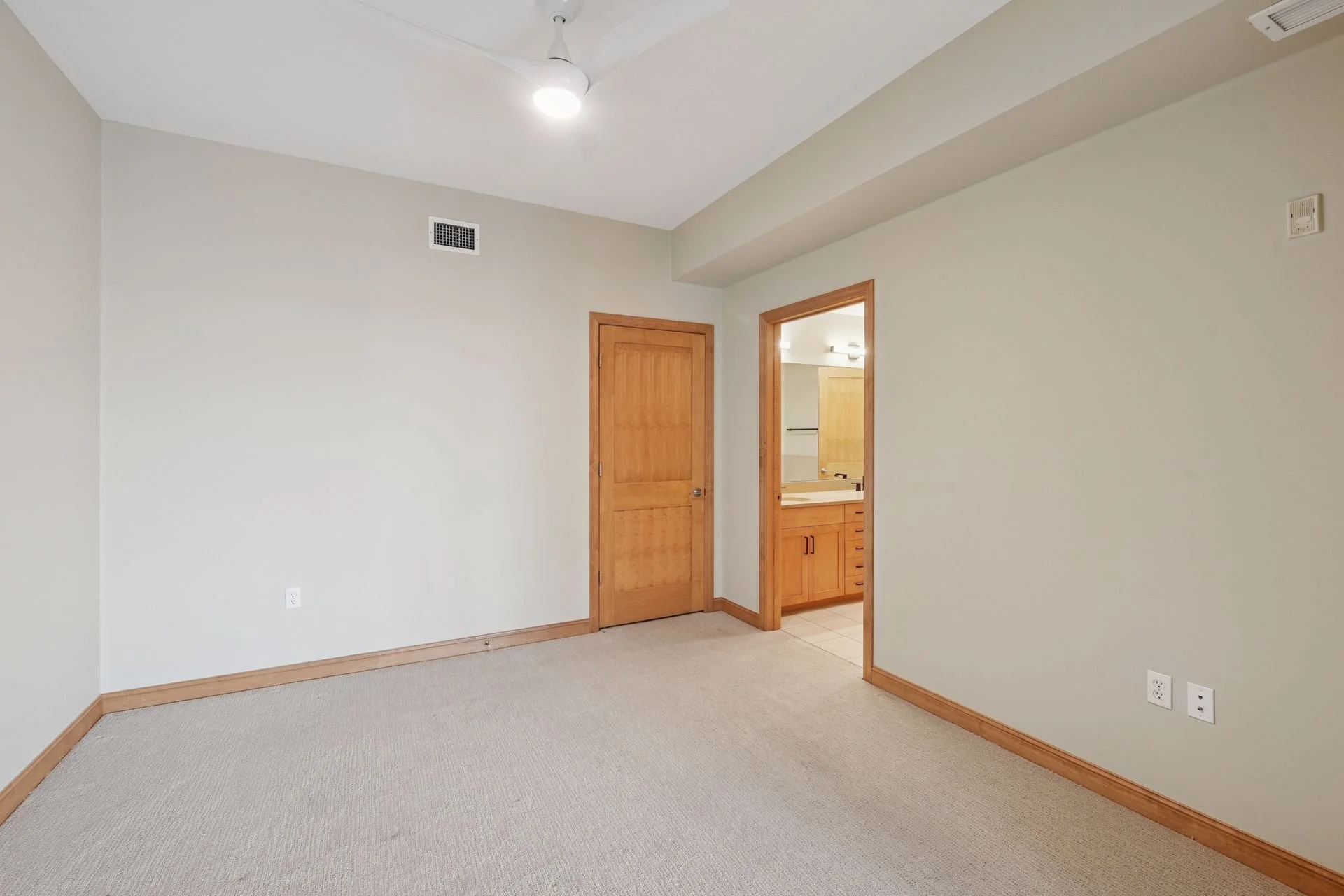 Empty beige room with wooden doorways and trim, carpeted floor, ceiling fan, and visible bathroom with wooden cabinets and sink.
