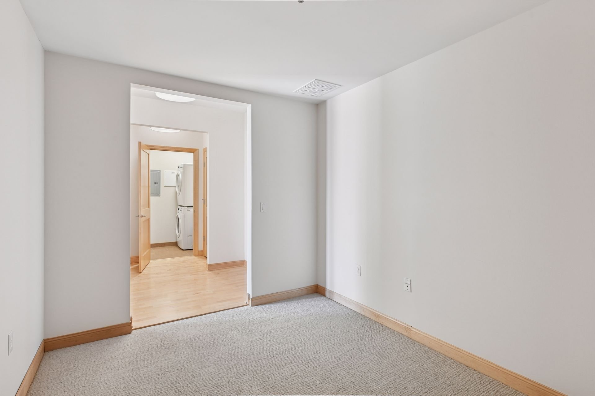 Empty room with light-colored carpet, white walls, wooden baseboards, and a doorway leading to a laundry area with stacked washer and dryer.