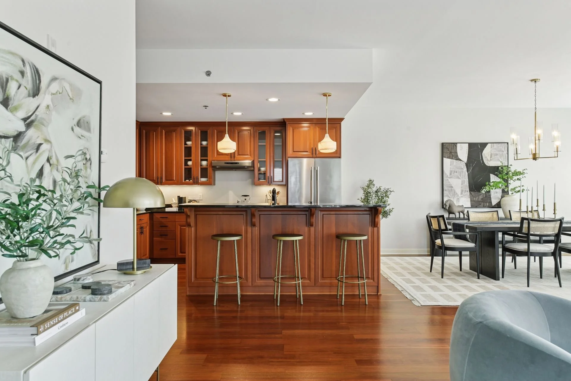 Open-concept kitchen and dining area with white walls and wooden floors, featuring a kitchen with wooden cabinets, stainless steel refrigerator, black countertop. Adjacent dining space has a chandelier.