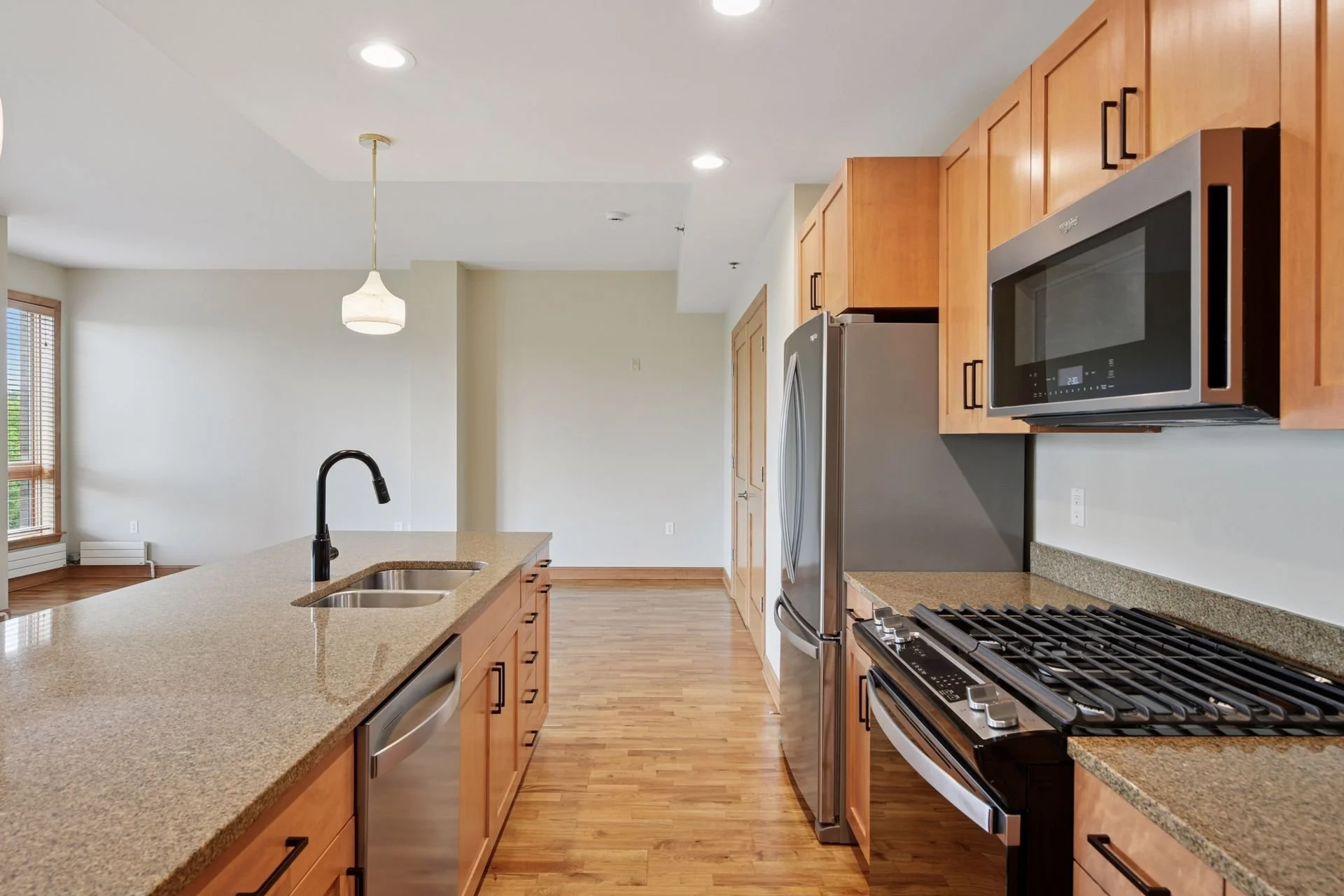 Modern kitchen with wooden cabinets, stainless steel appliances, a granite countertop island with a sink, and hardwood floors.