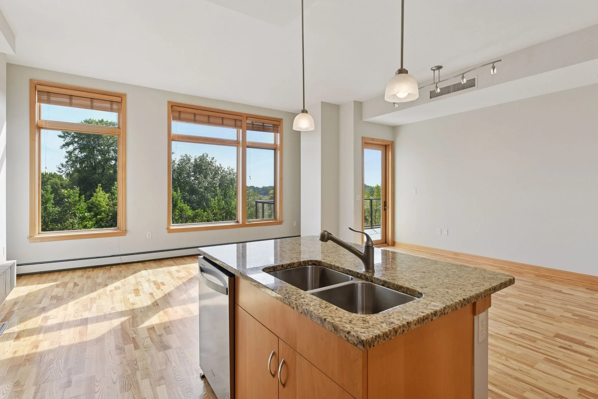 Empty modern kitchen and living area with large windows, hardwood floors, granite island, and a balcony door.